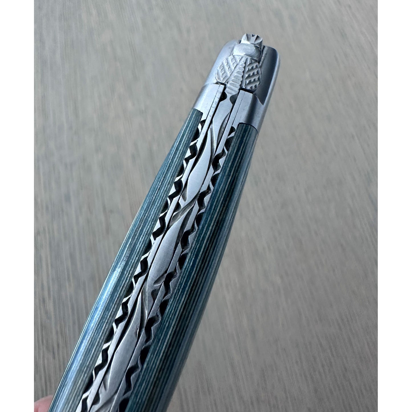 A close-up of a folding knife shown diagonally, highlighting its metallic silver patterned blade and reddish-brown textured handle against a light gray wood-grain background.