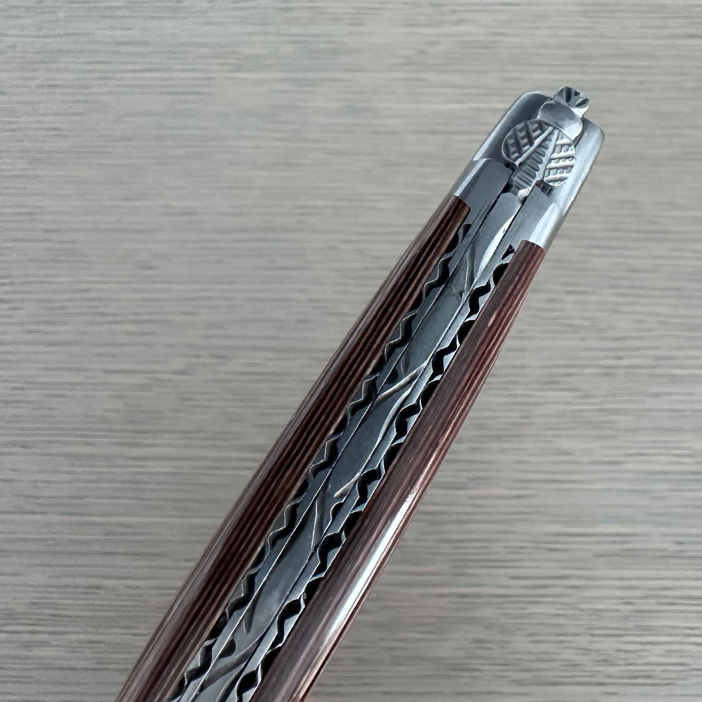 A close-up of a folding knife shown diagonally, highlighting its metallic silver patterned blade and reddish-brown textured handle against a light gray wood-grain background.
