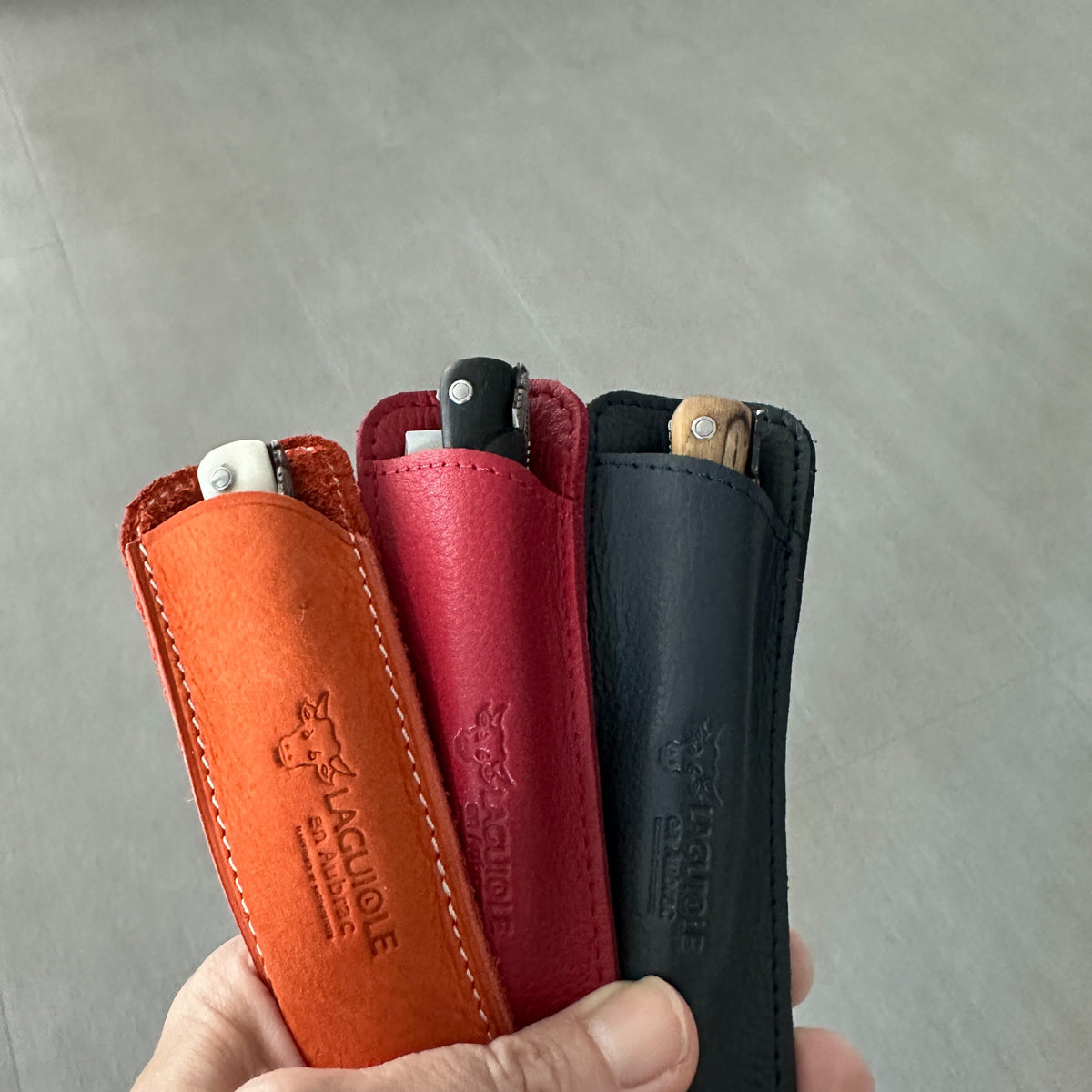 Three leather knife sheaths in orange, red, and marine, each holding a folding knife, held in a hand against a gray concrete background. The sheaths are embossed with the brand name Laguiole en Aubrac.