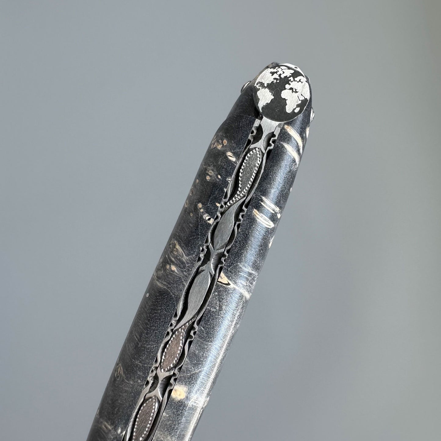 A close-up of a dark folding knife, ornate filigree detailing and a white textured tip, set against a solid gray background.