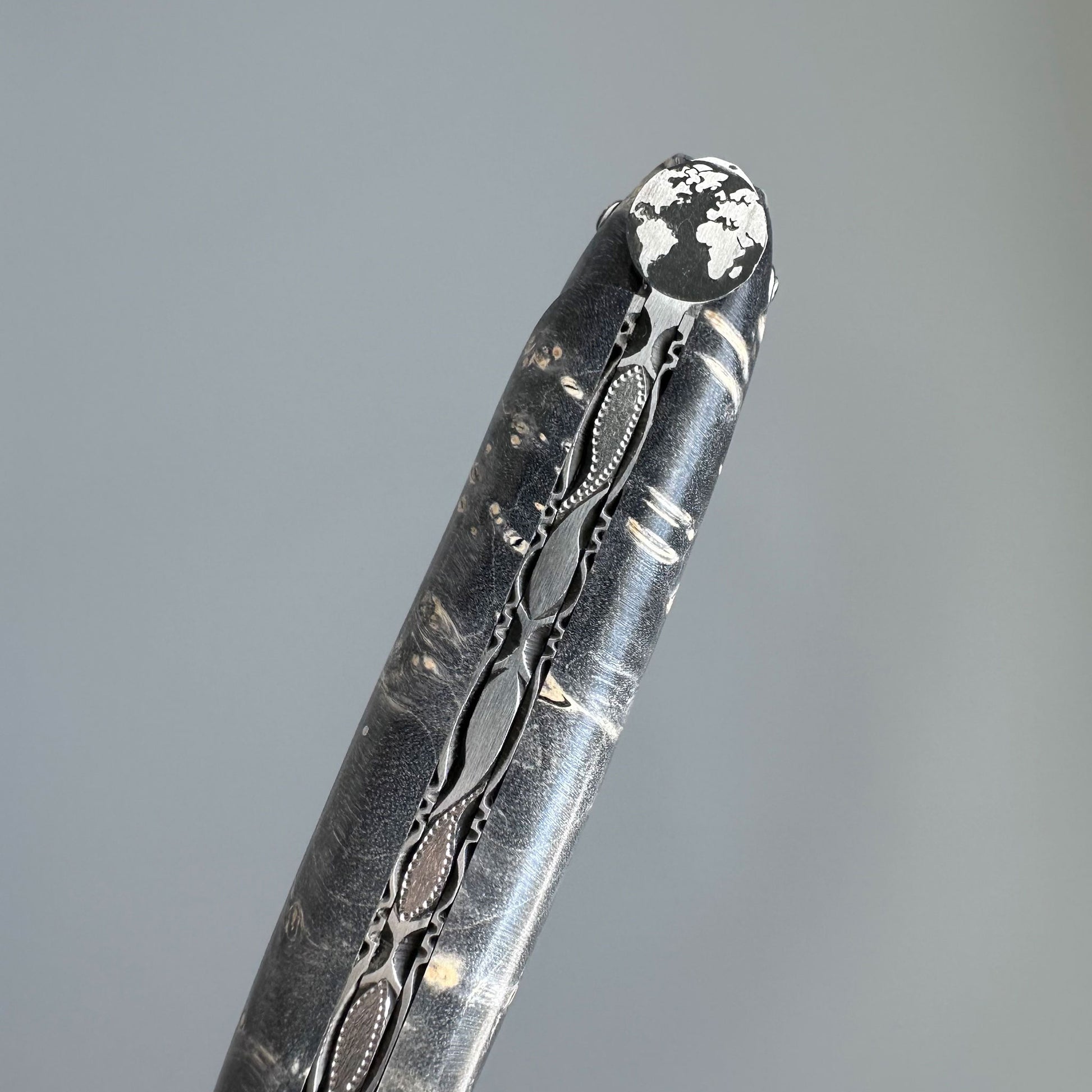 A close-up of a dark folding knife, ornate filigree detailing and a white textured tip, set against a solid gray background.