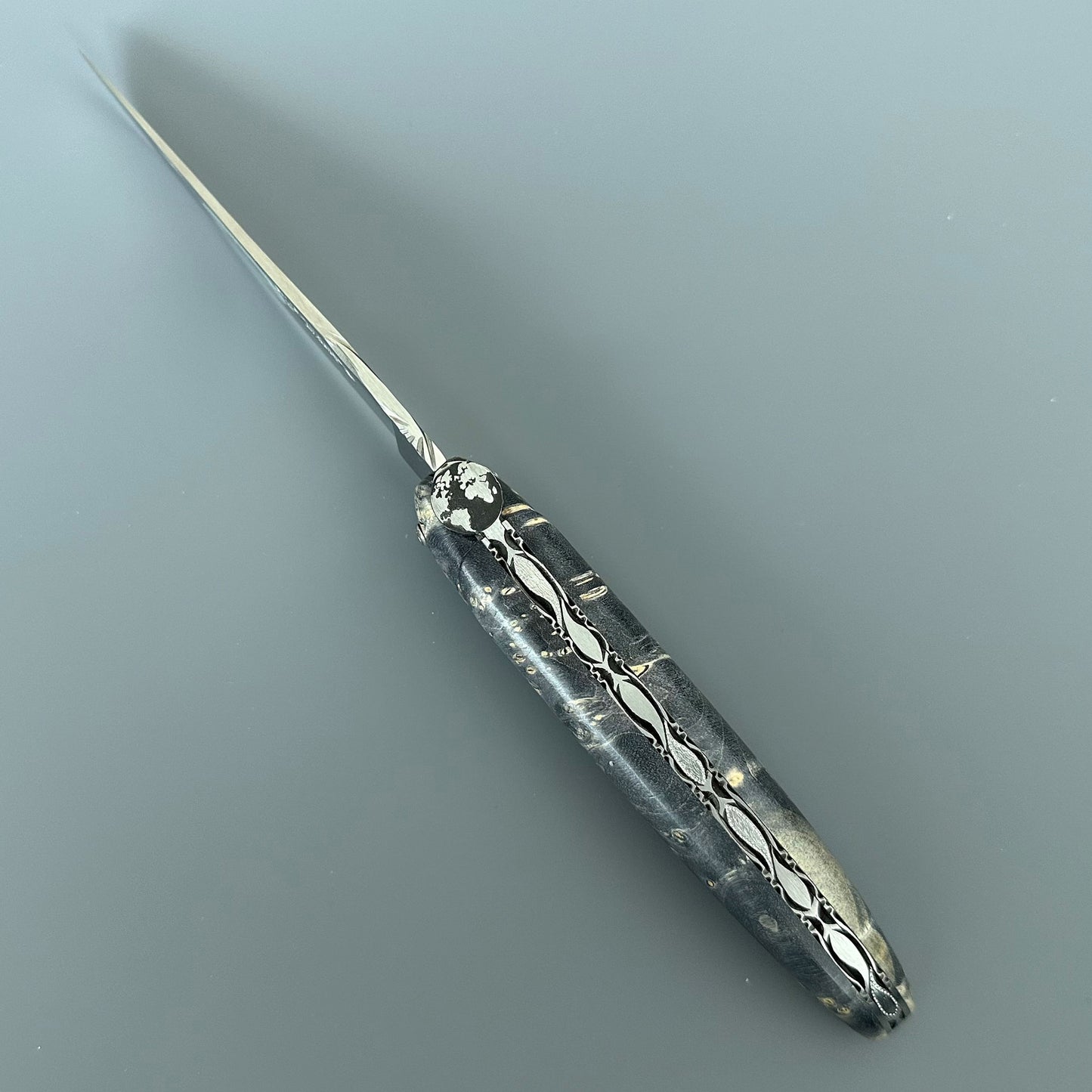 Folding pocket knife with an ornate, decorative handle and a long, slender blade, photographed on a smooth gray surface.