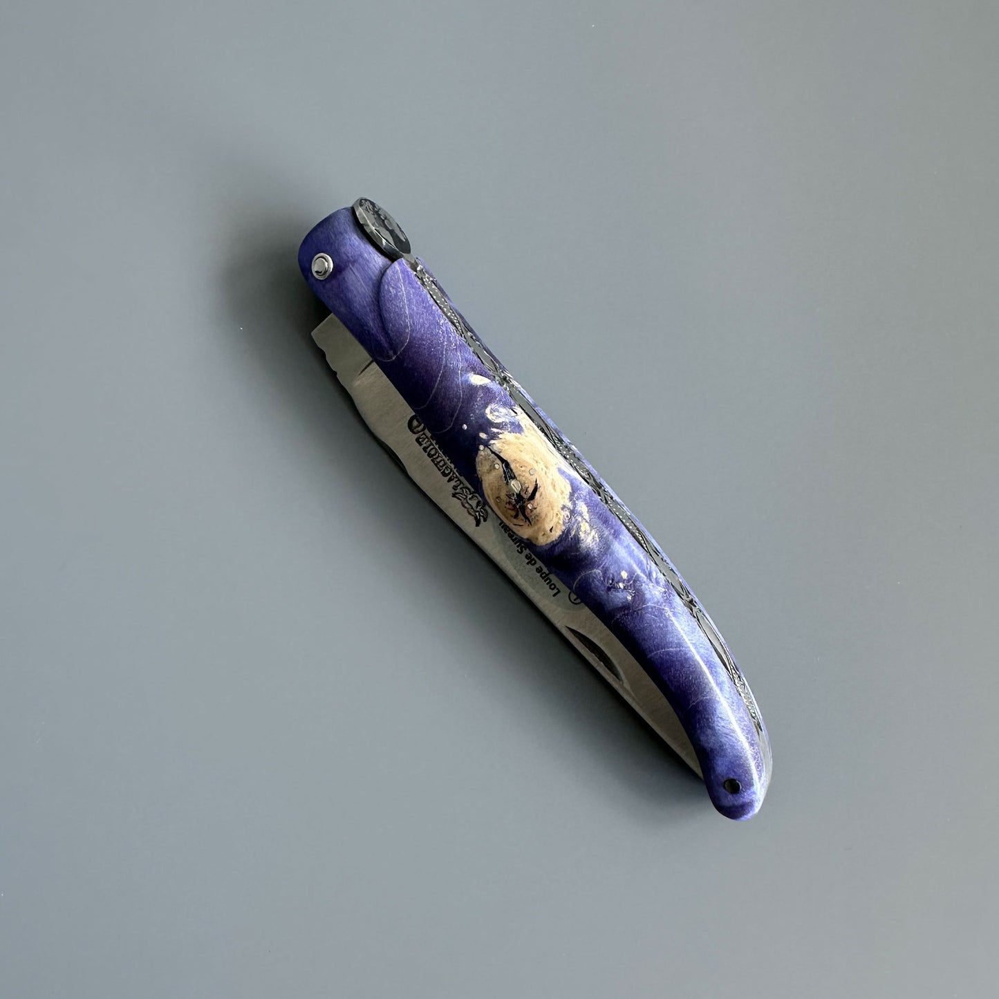 A folding pocket knife with a purple and blue marbled handle and a gold-toned moon design, shown open on a smooth gray surface.