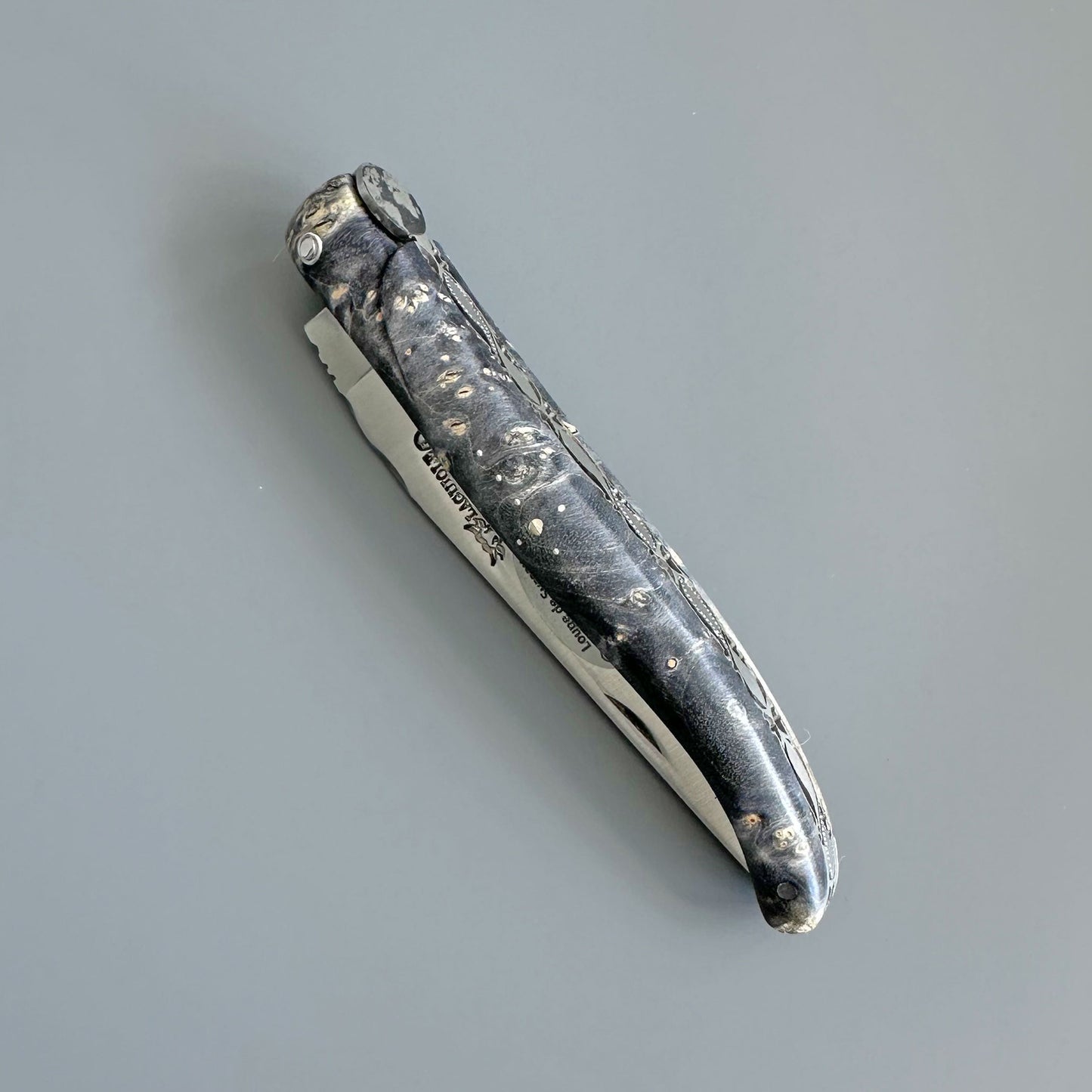 A folding pocket knife with a textured black handle featuring white marbled or burl-like patterns, and a silver metal blade partially extended, shown on a plain gray background.