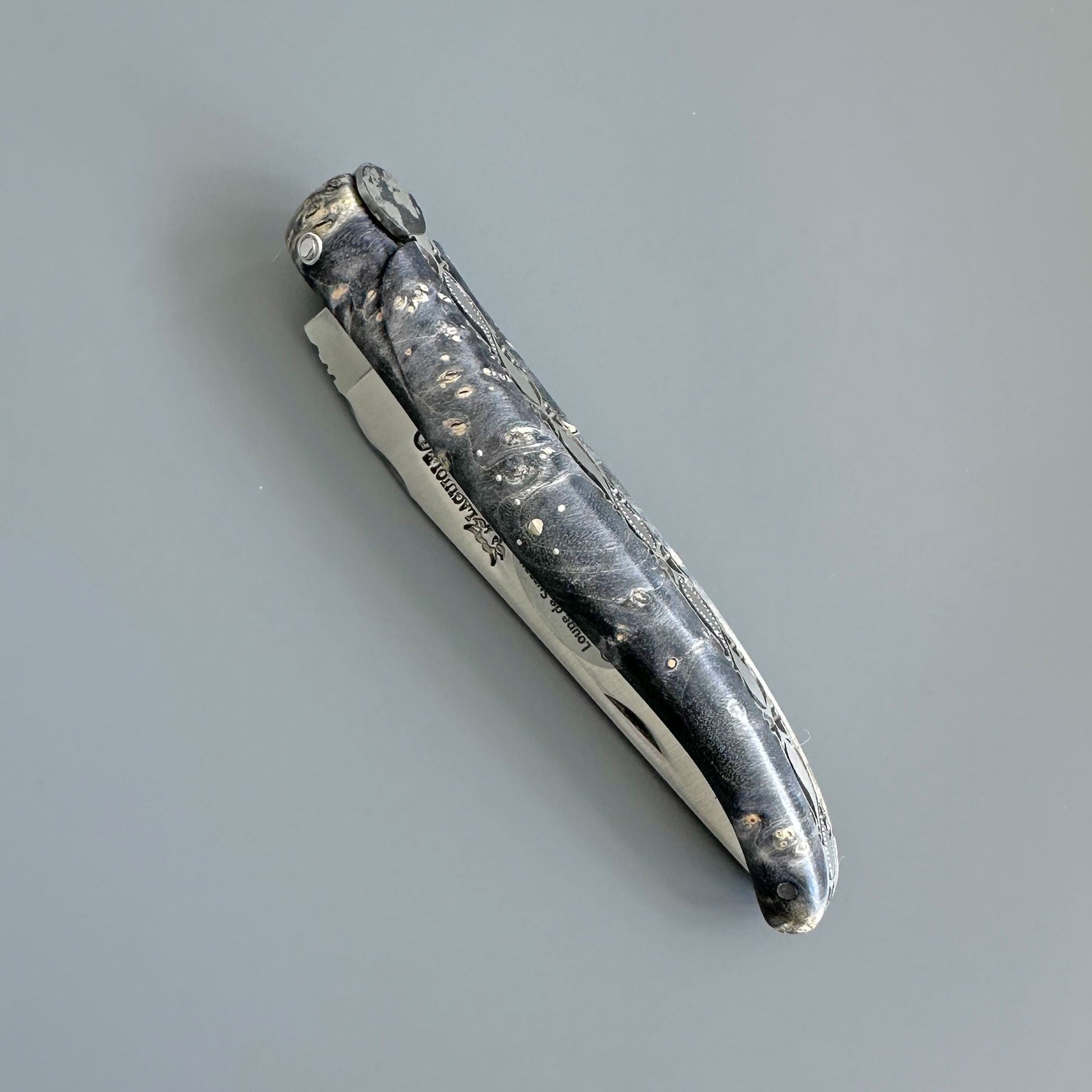 A folding pocket knife with a textured black handle featuring white marbled or burl-like patterns, and a silver metal blade partially extended, shown on a plain gray background.