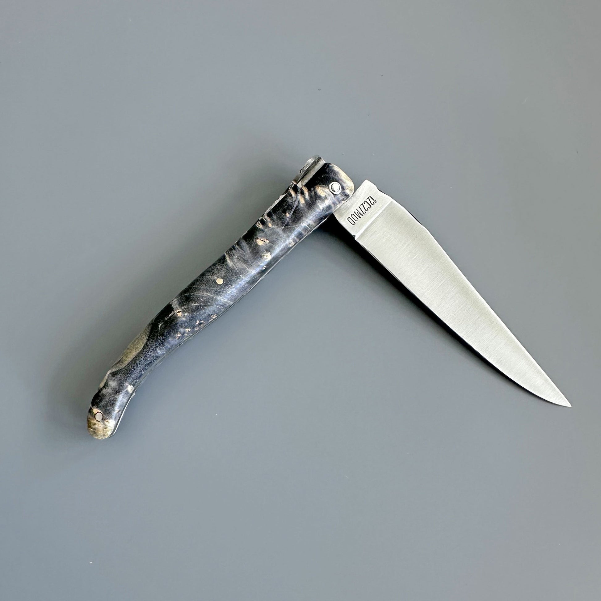 Laguiole en Aubrac Cosmos folding pocket knife with a textured black and gray handle and a silver stainless steel blade, shown open on a solid gray background. 