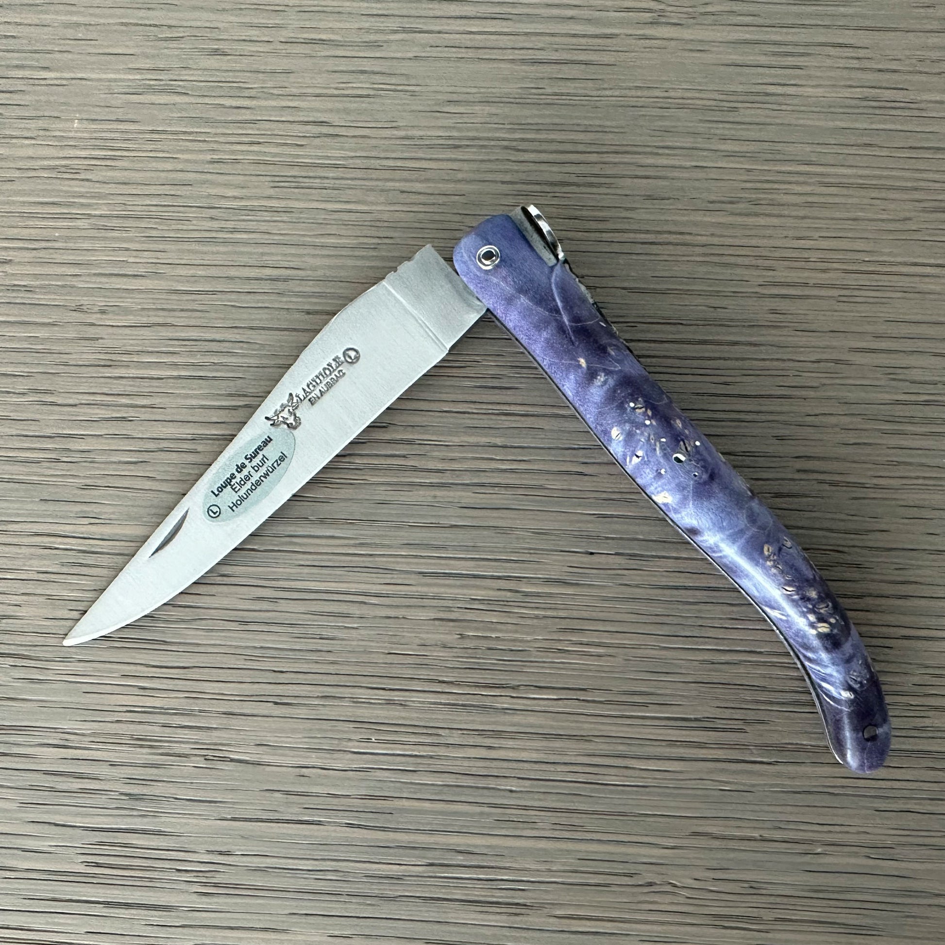 Laguiole en Aubrac Cosmos purple marbled handle, shown open on a gray wood-grain surface. 