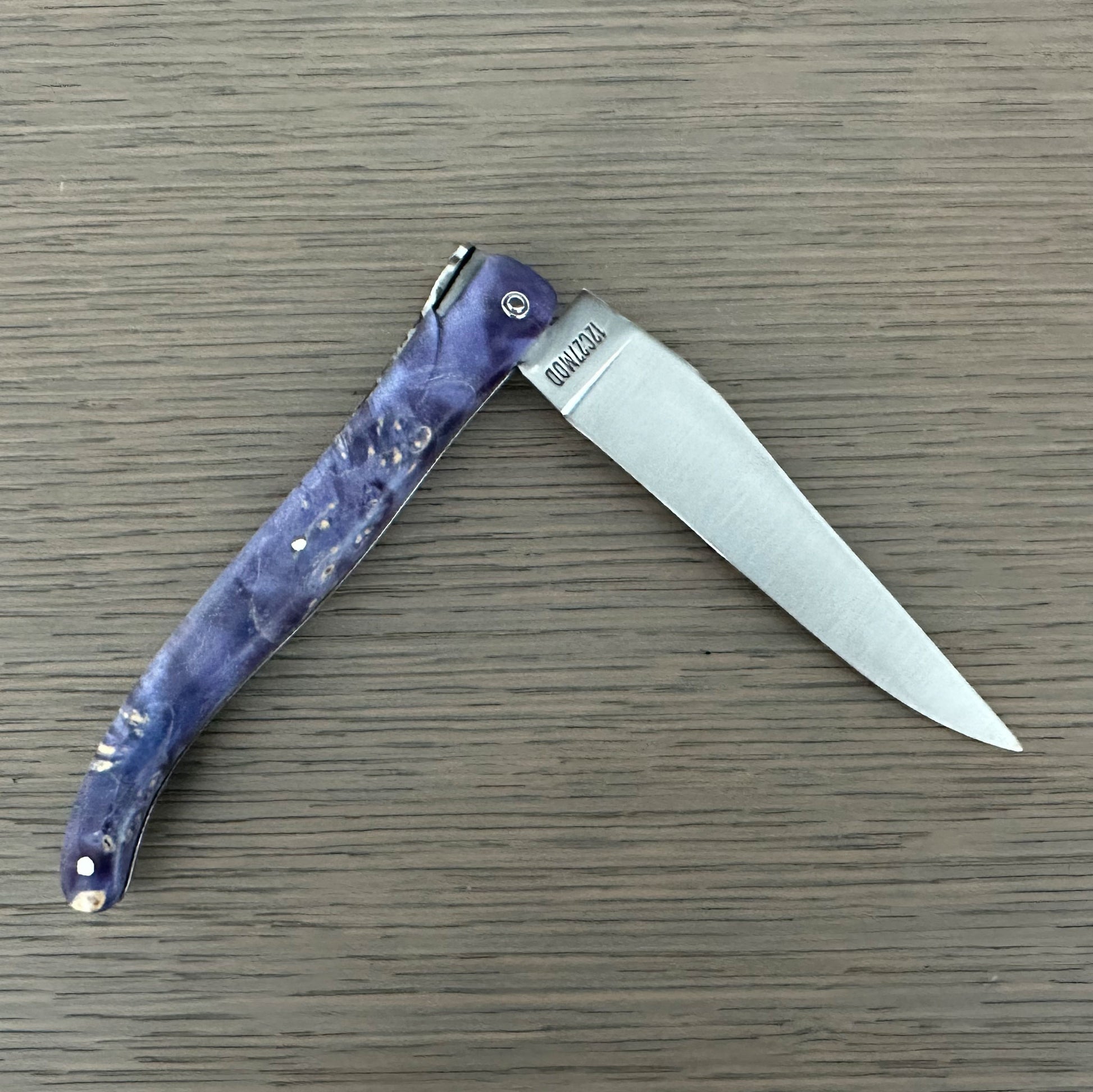 Laguiole en Aubrac folding knife with textured purple handle and a silver stainless steel blade, shown open on a gray wood-grain surface. 