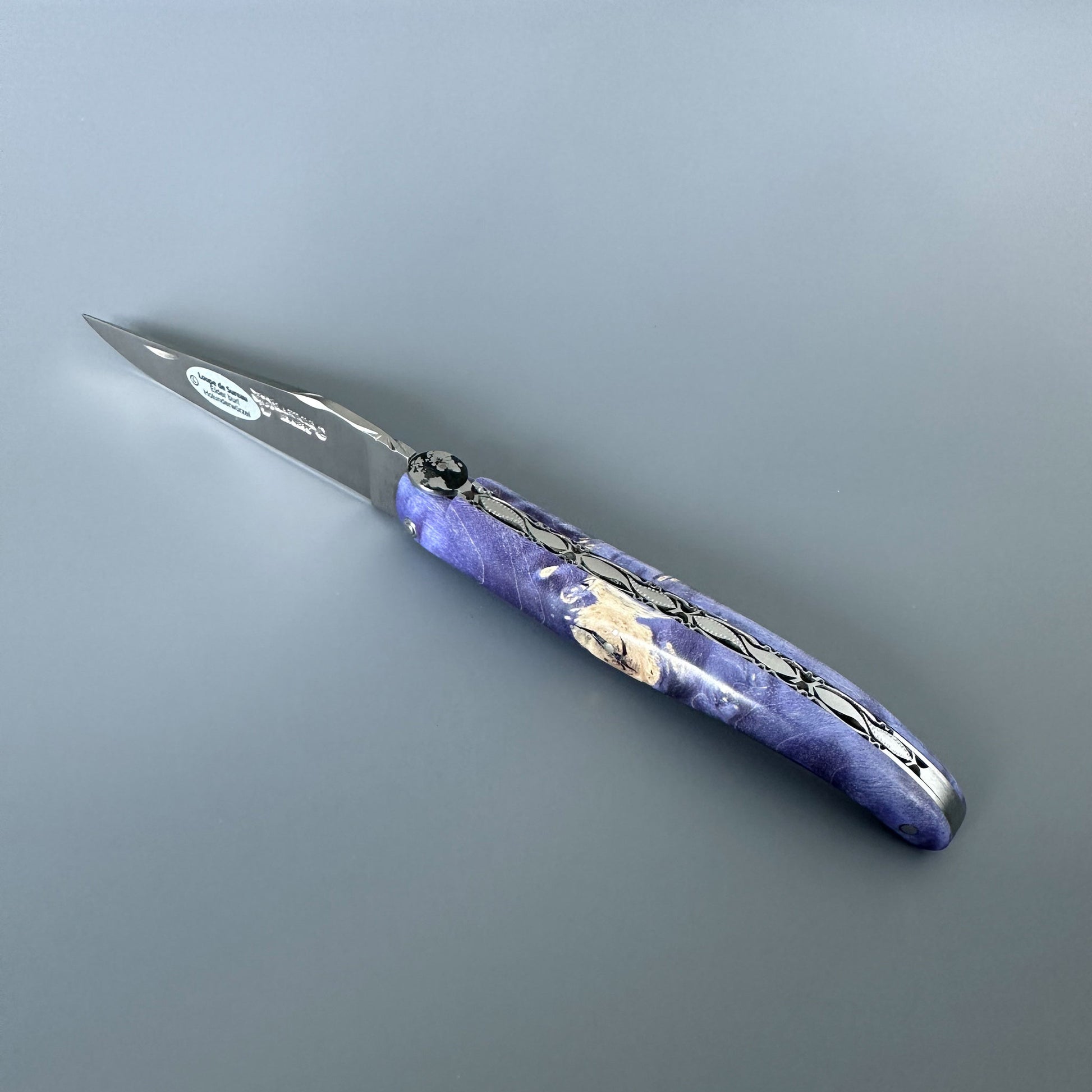 Laguiole en Aubrac Csomos folding pocket knife with Earth bee, stainless steel blade and a purple marbled handle featuring a cream tone shown on a neutral gray background.