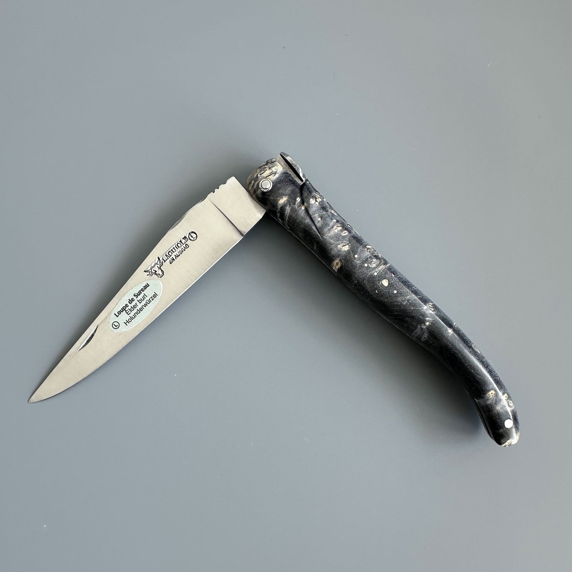 Laguiole en Aubrac folding pocket knife shown open on a solid gray background. The blade is engraved and handle is made of might Earl Burl