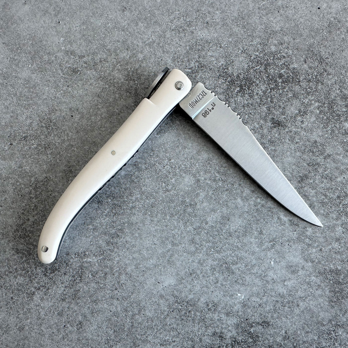 Laguiole en Aubrac pocket knife with Ivory handle and a silver steel blade, shown open on a textured gray concrete surface. 