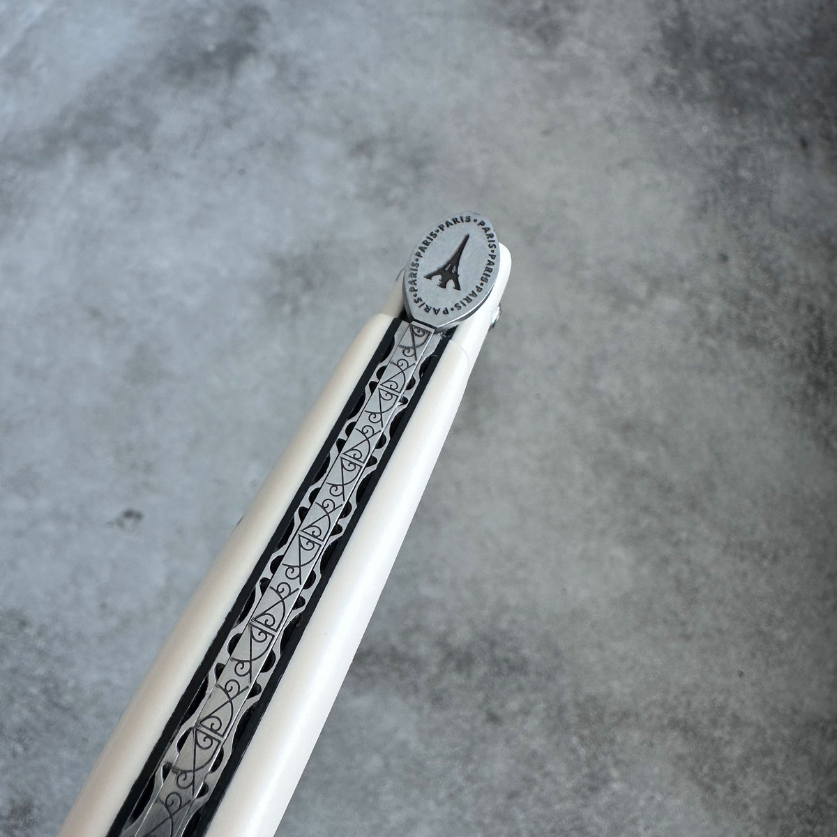 A close-up of a Laguiole en Aubrac silver Eiffel Tower emblem on the tip of the knife set against a textured gray concrete background.
