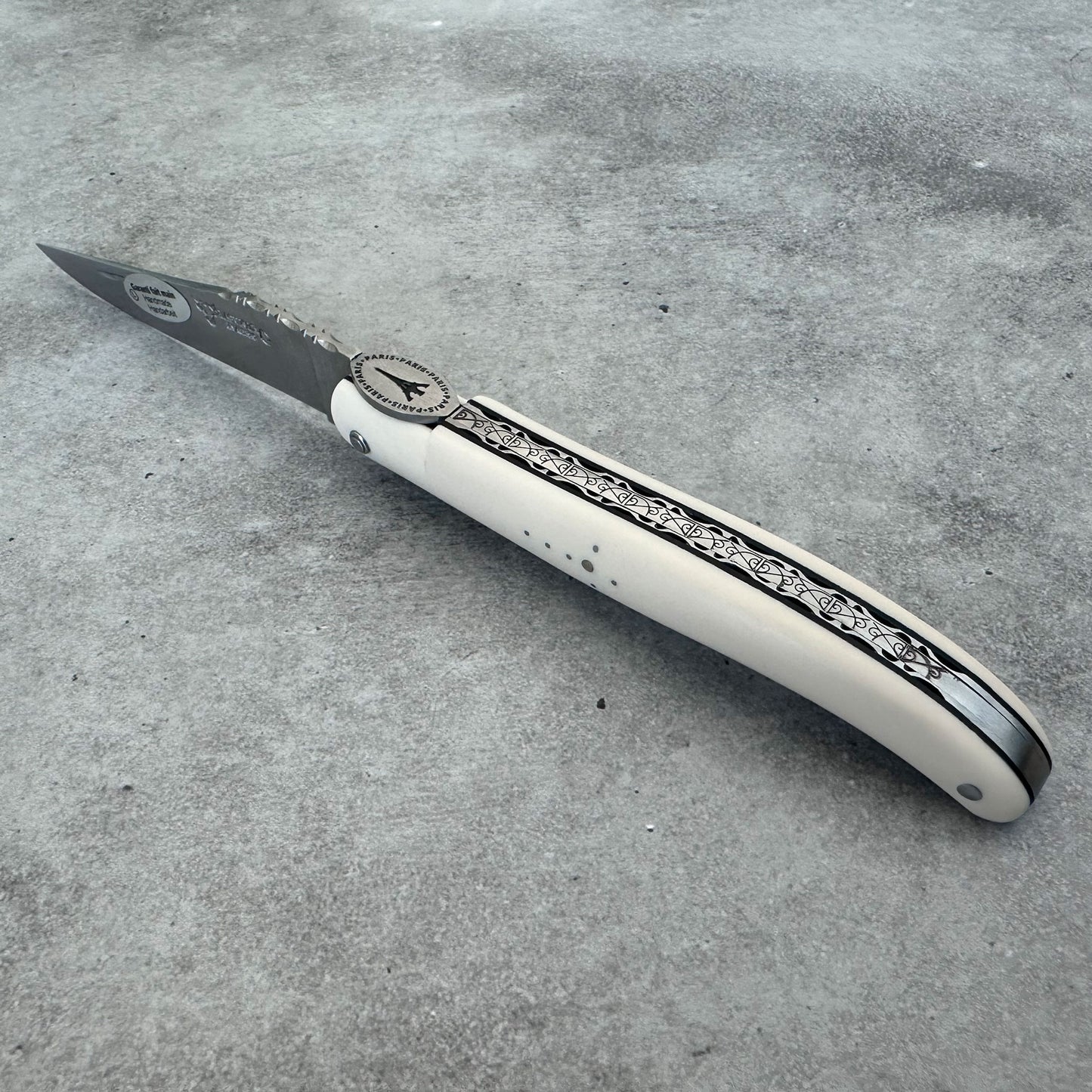 Laguiole en Aubrac Eiffel knife with a ivory colored handle and a black engraved pattern, shown on a textured gray concrete surface. 