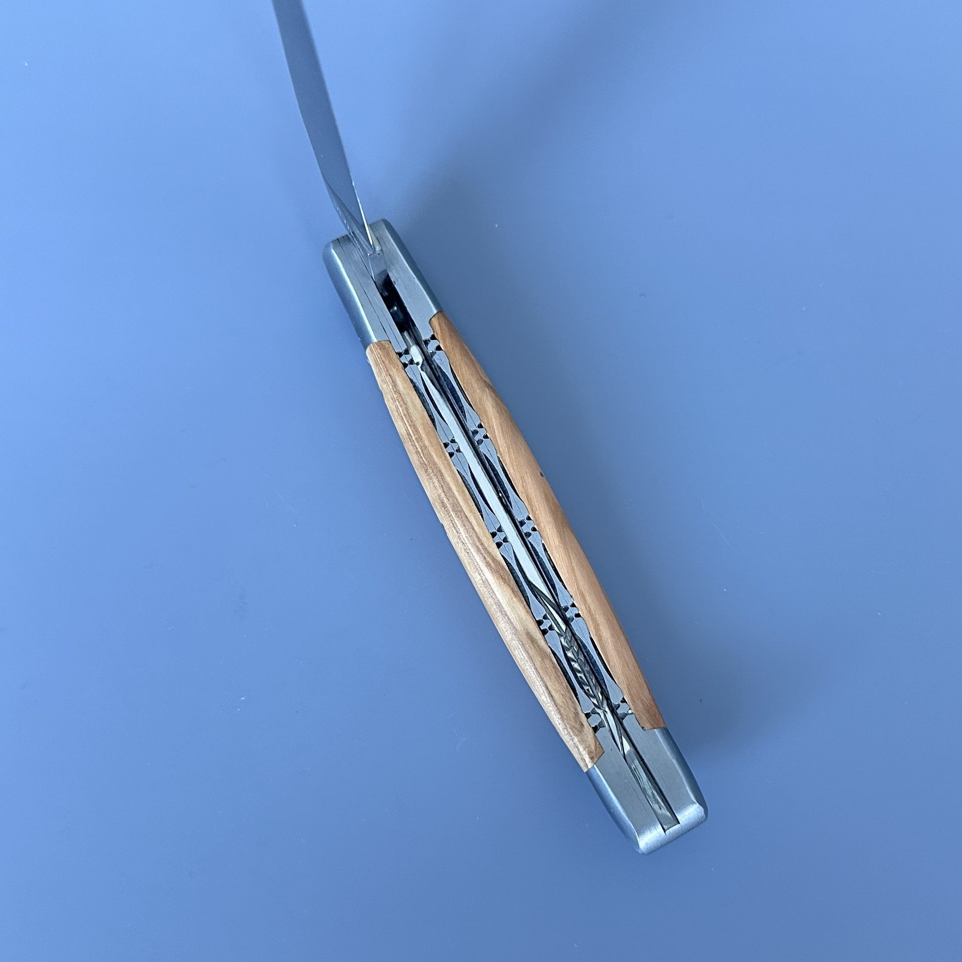 Inner spine of Laguiole Double Plated folding pocket knife with a light wood handle and silver metal blades, shown partially open on a solid blue background.