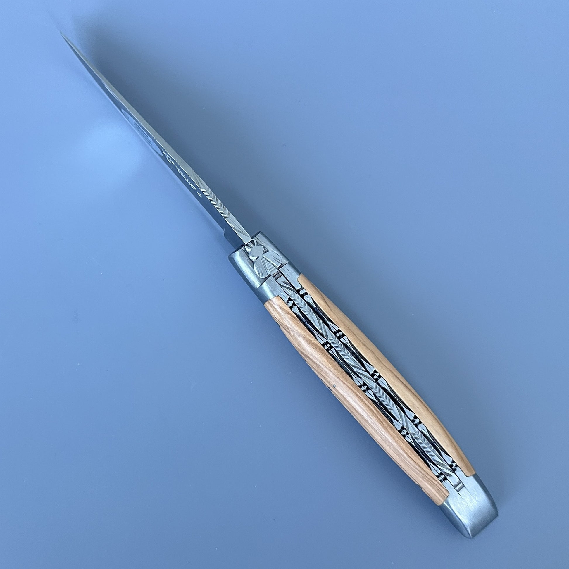 Cenital view of Laguiole Double Plated folding knife fully open, showing the polished blade, engraved bee bolster, and olive wood handle with decorative filework.