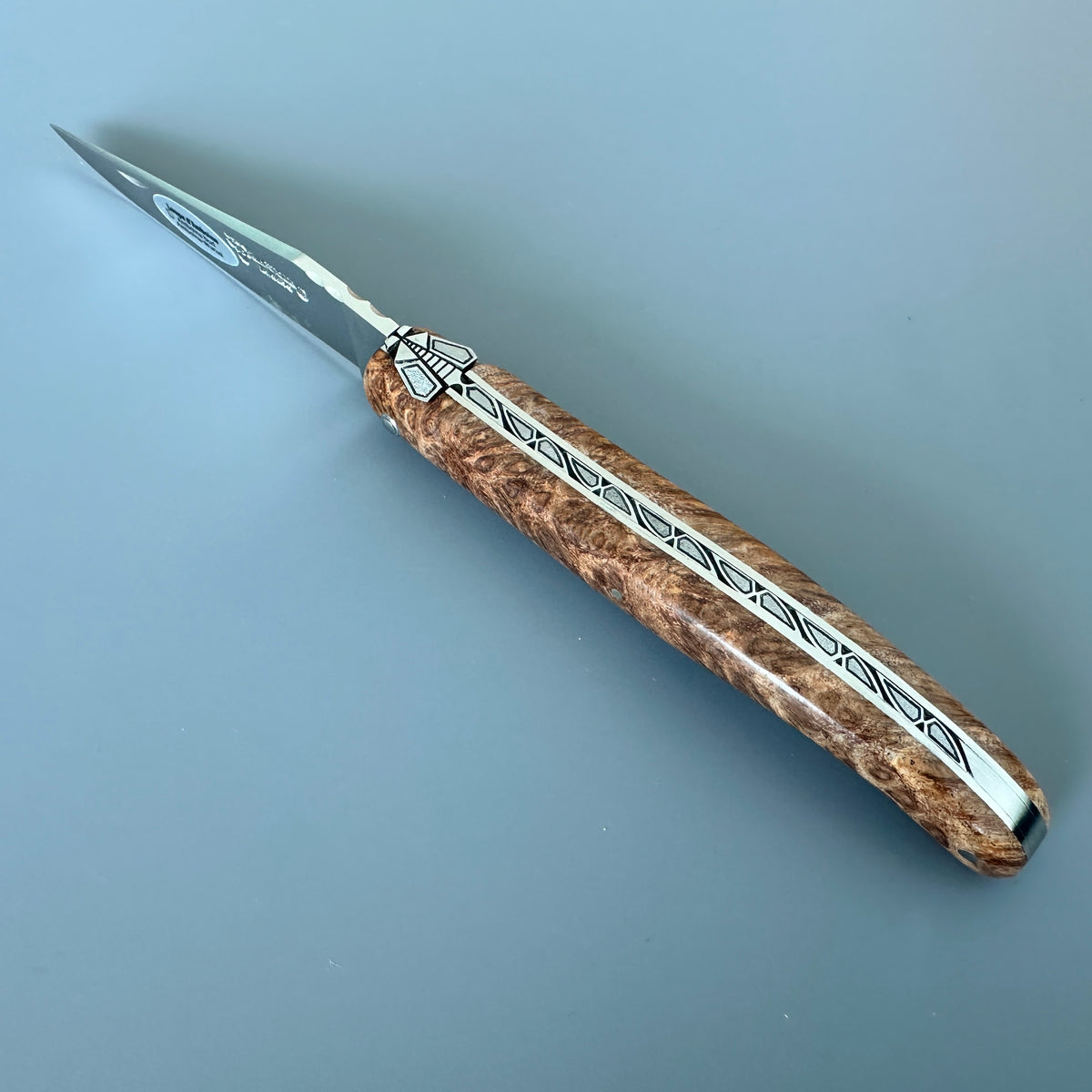 Laguiole en Aubrac folding knife with Amboyna burl wood handle featuring a central silver inlay with black geometric patterns, set against a solid blue-gray background.