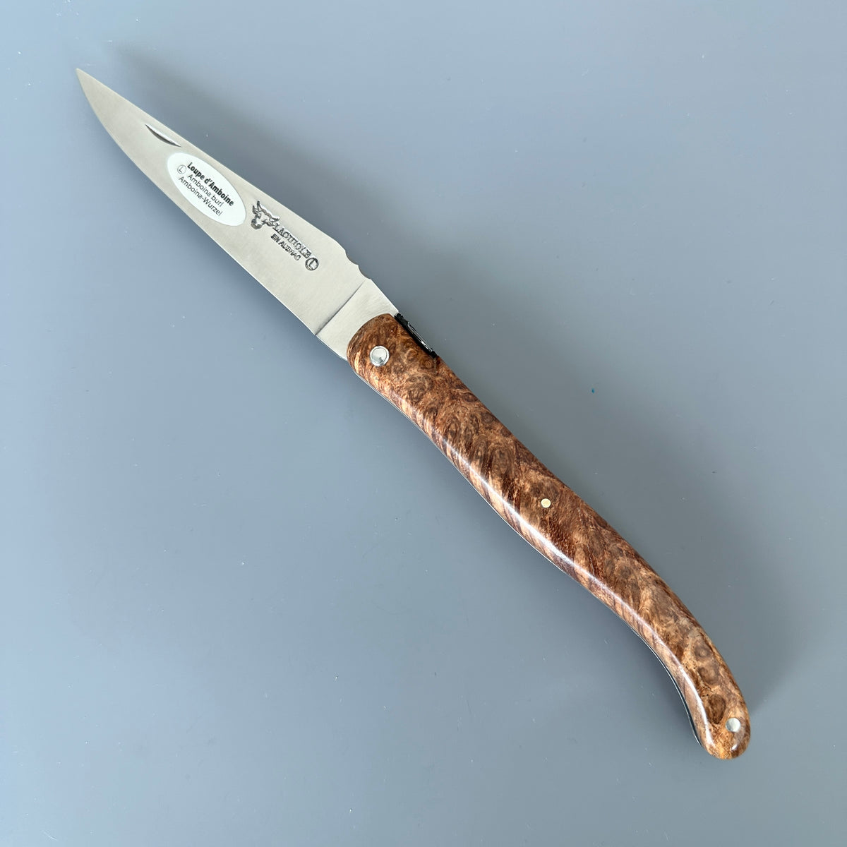 Laguiole en Aubrac knife with richly textured Amboyna burl wood handle, displayed diagonally on a smooth gray background.
