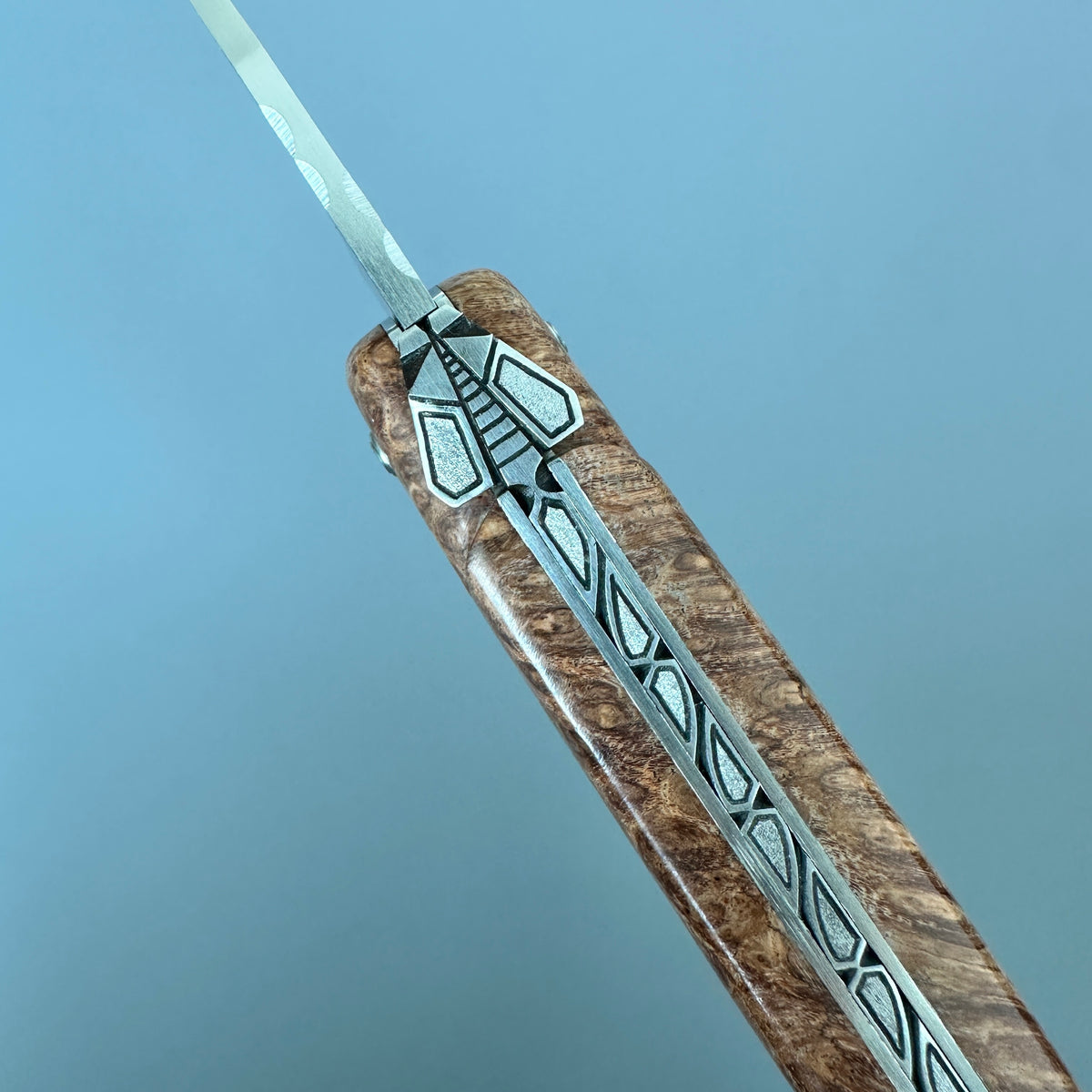 Laguiole en Aubrac Folding Knife with Amboyna burl close-up of a folding knife featuring geometric inlay details, set against a solid blue background.