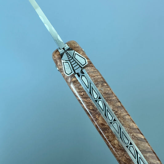 Laguiole en Aubrac Folding Knife with Amboyna burl close-up of a folding knife featuring geometric inlay details, set against a solid blue background.