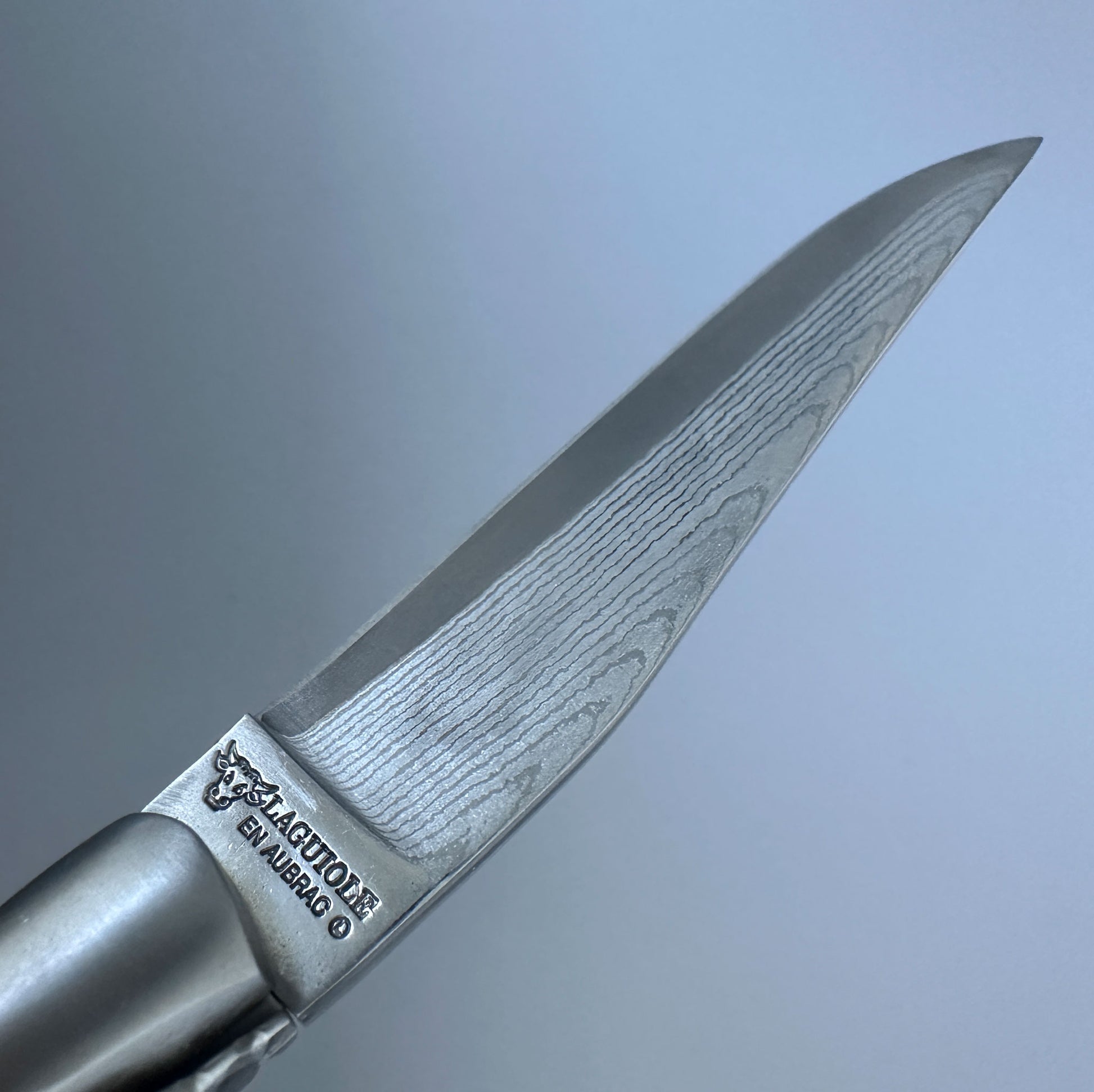 A close-up of a Damascus steel knife blade with a visible wavy pattern, featuring the brand engraving 'Laguiole en Aubrac' on the bolster, set against a soft gray gradient background.