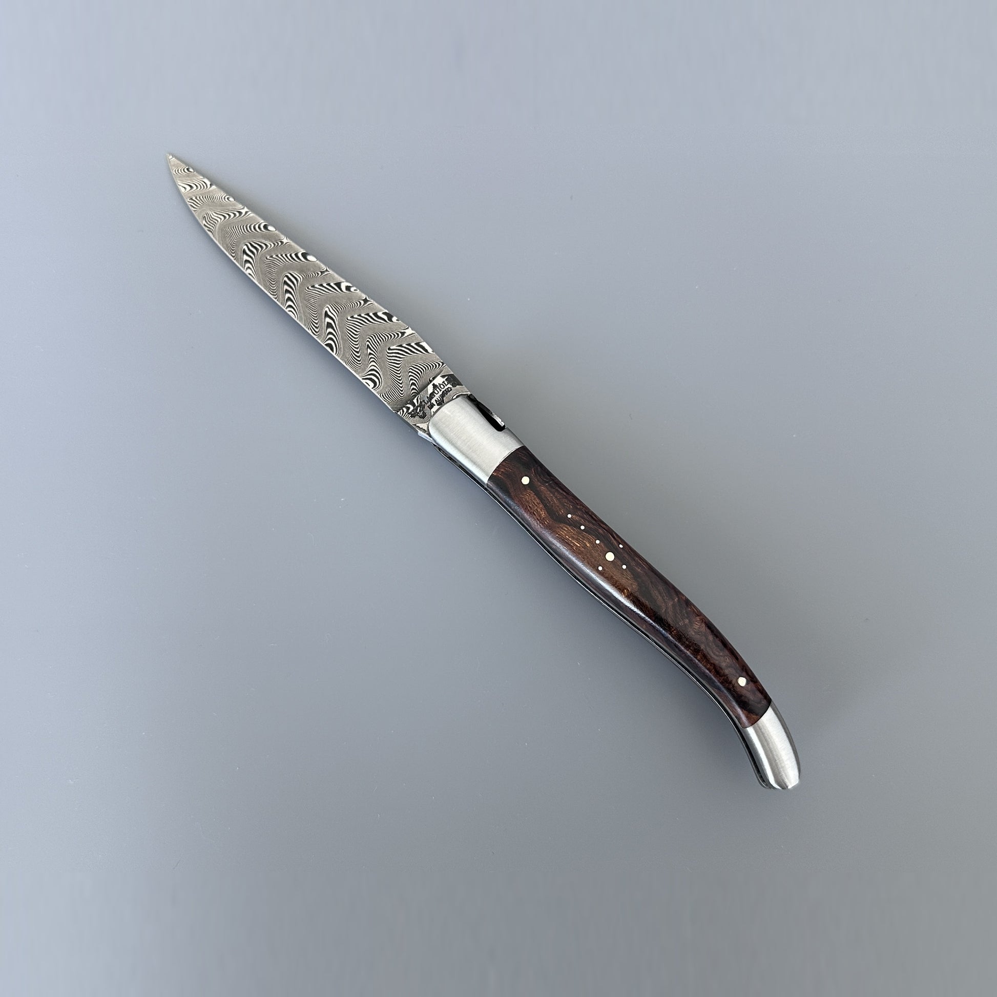 A folding pocket knife with a Damascus steel blade and a dark brown wood handle, shown diagonally on a neutral gray background.
