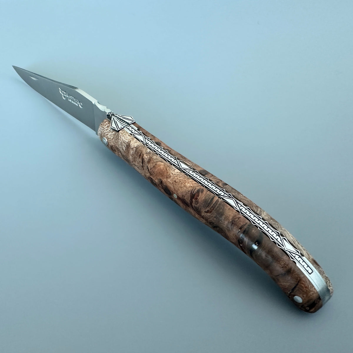 Laguiole en Aubrac folding pocket knife with satin blade and Albizia burl wood handle, shown on a solid light blue-gray background.