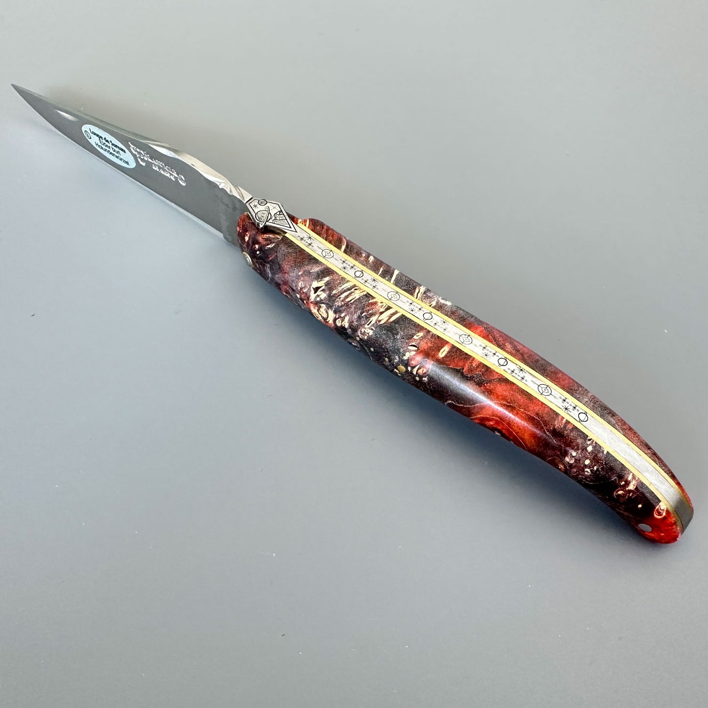 A folding pocket knife with a tinted poplar burl decorative handle made of red, dark brown and gold marbled material, accented by a gold center stripe. 