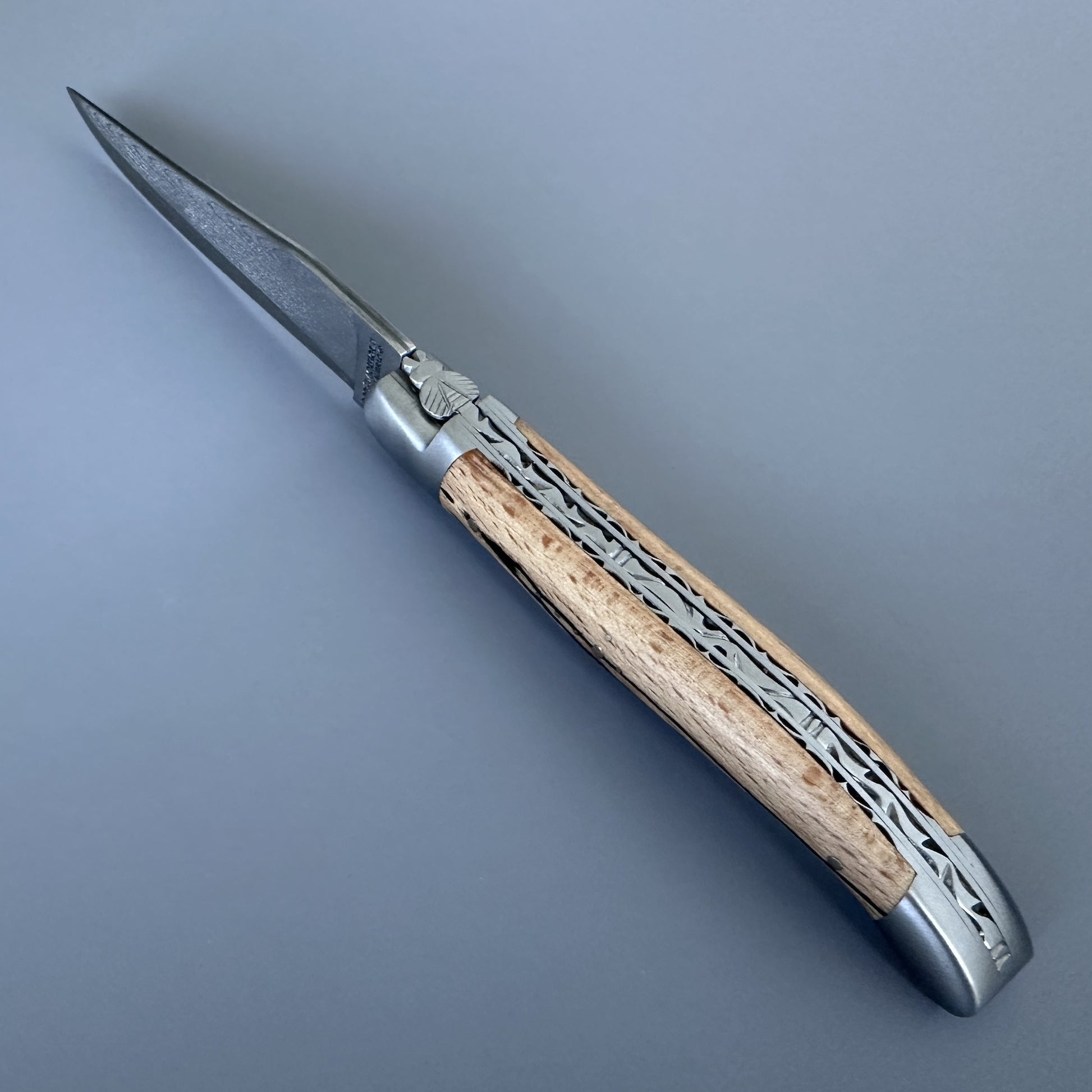 Laguiole Double Plate Damascus folding knife with Aubrac wood handle, blade open, on a blue background.