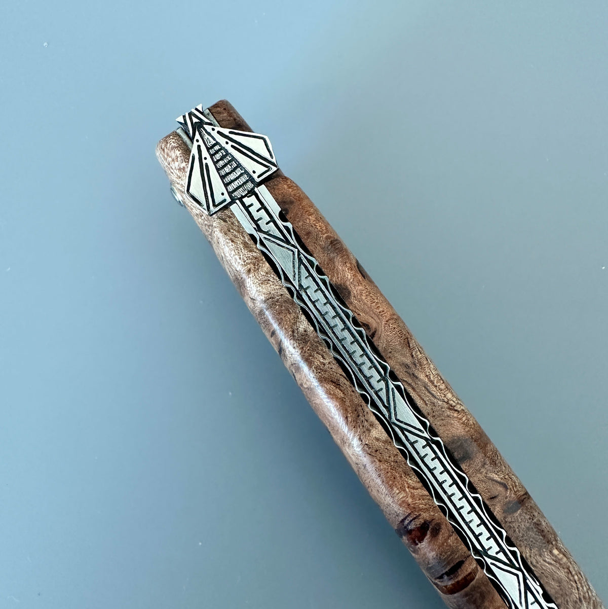 A close-up of a Laguiole en Aubrac Belle Piece with intricate geometric inlay patterns, set against a solid light blue background.