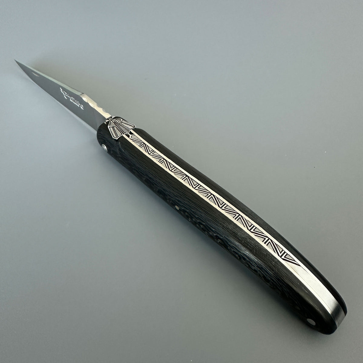 Open Laguiole en Aubrac Belle Piece Carbon folding knife showing its blade, patterned handle, geometric bee and engraved spine.