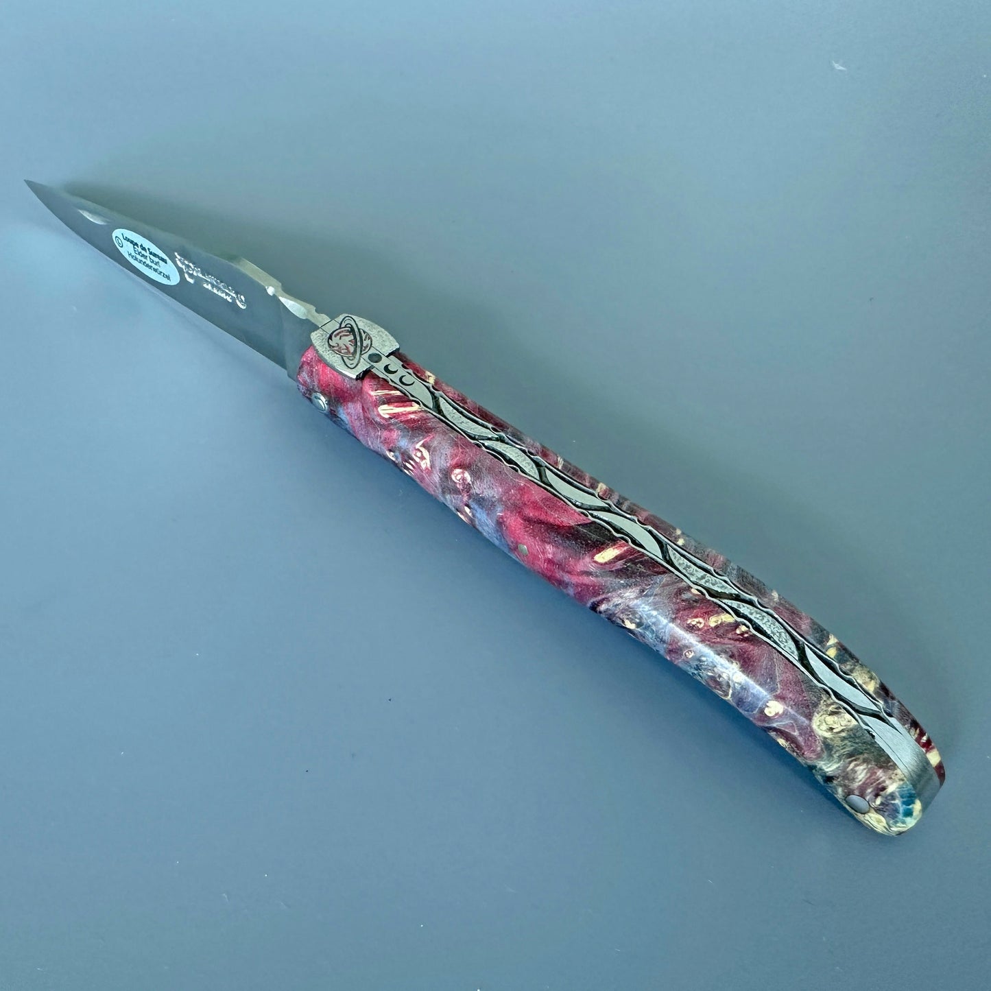 A folding pocket knife with a dark gray steel blade and a highly decorative, marbled handle featuring pink, purple, blue tones, set against a solid slate gray background.