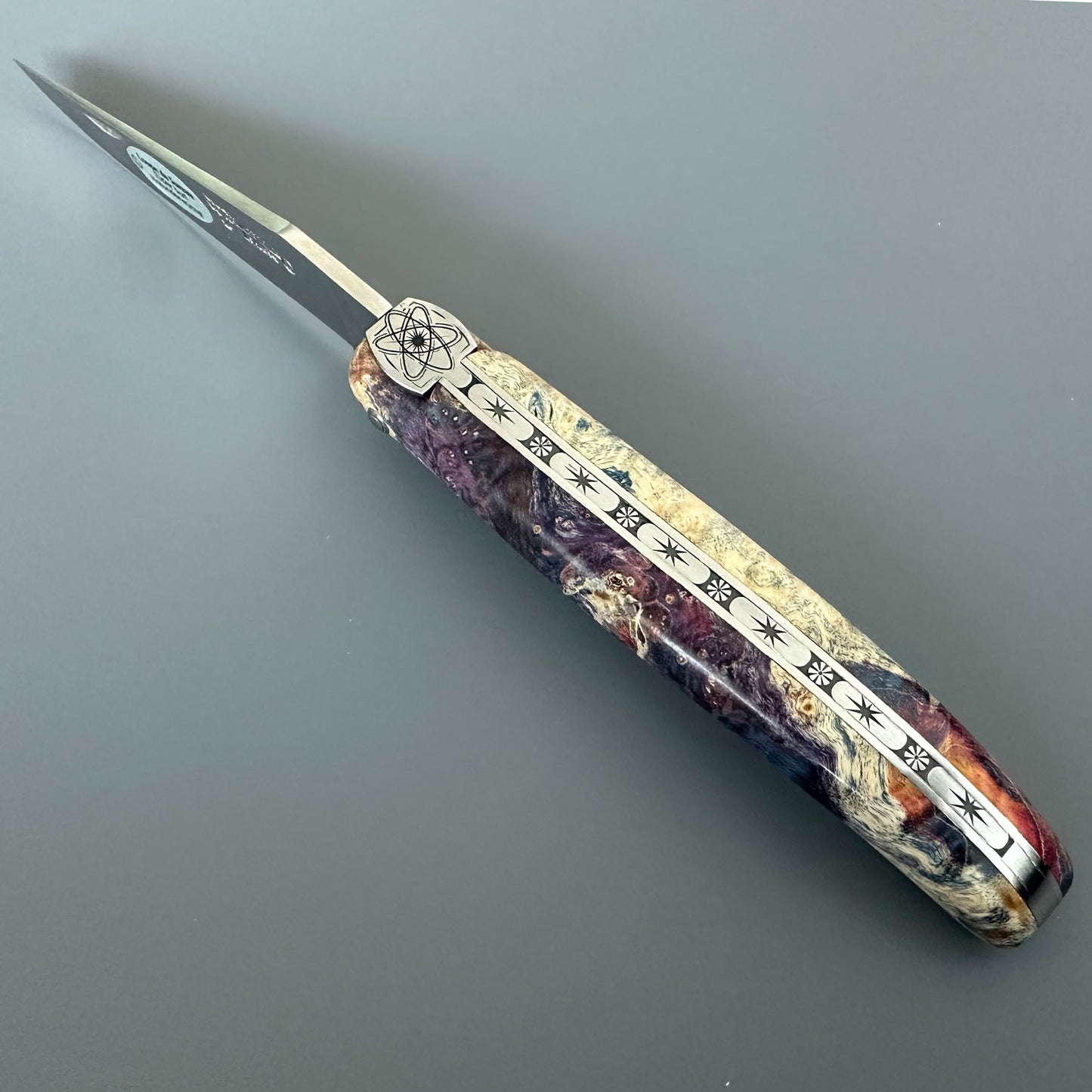 A folding pocket knife with a silver blade and a highly detailed, multicolored handle. The handle features a cream-colored base inlaid with purple, red, and orange marbled patterns, and engraved with silver symbols and text.
