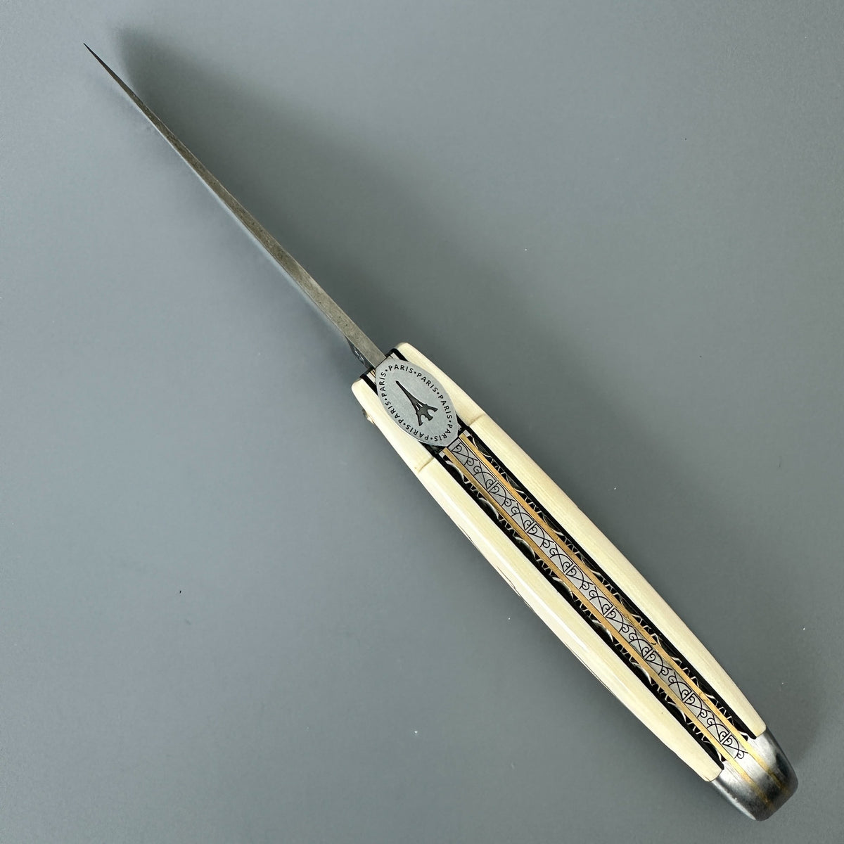 Laguiole en Aubrac folding pocket knife with a damascus blade and a mammoth ivory crust featuring black and gold inlay patterns over a gray background