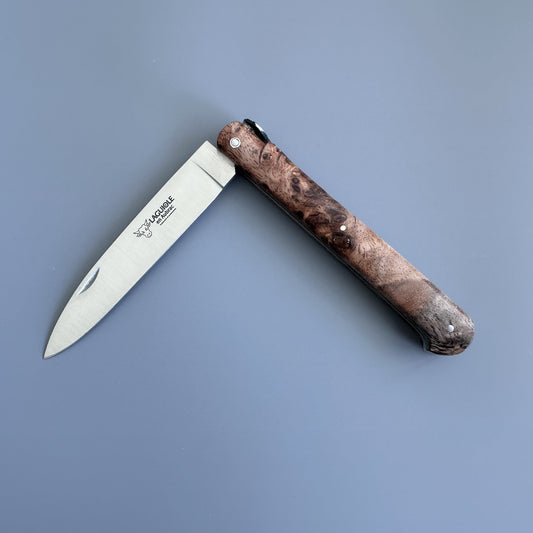 A folding pocket knife with a silver steel blade and a dark brown burl wood handle, shown open on a solid blue-gray background. The blade features the brand name: Laguiole en Aubrac