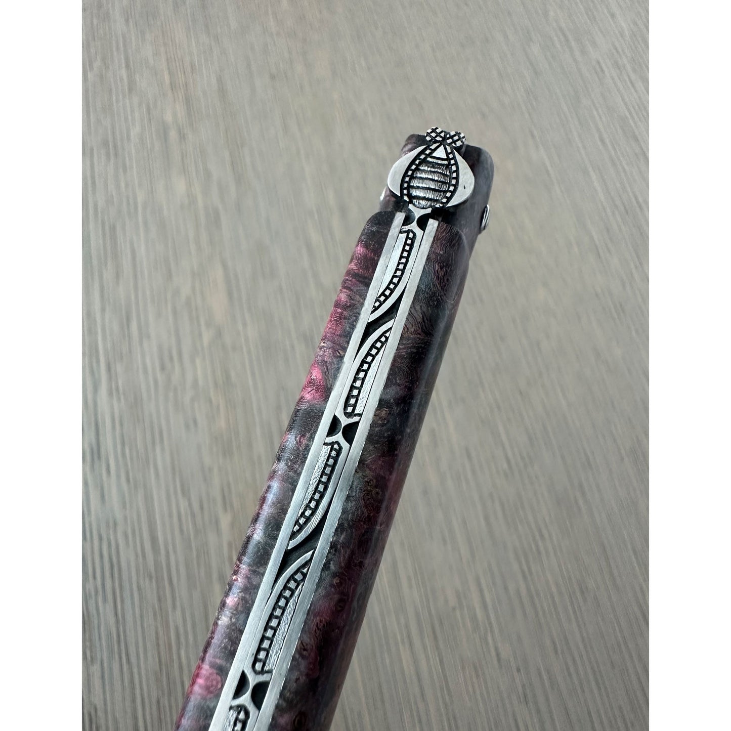 A close-up of bee handle with a marbled dark purple and pink resin body, featuring a silver-colored metal inlay with black geometric patterns and a silver pommel.