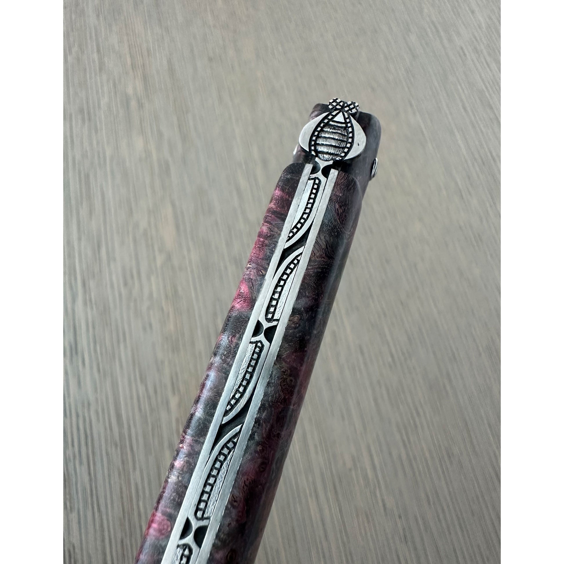 A close-up of bee handle with a marbled dark purple and pink resin body, featuring a silver-colored metal inlay with black geometric patterns and a silver pommel.
