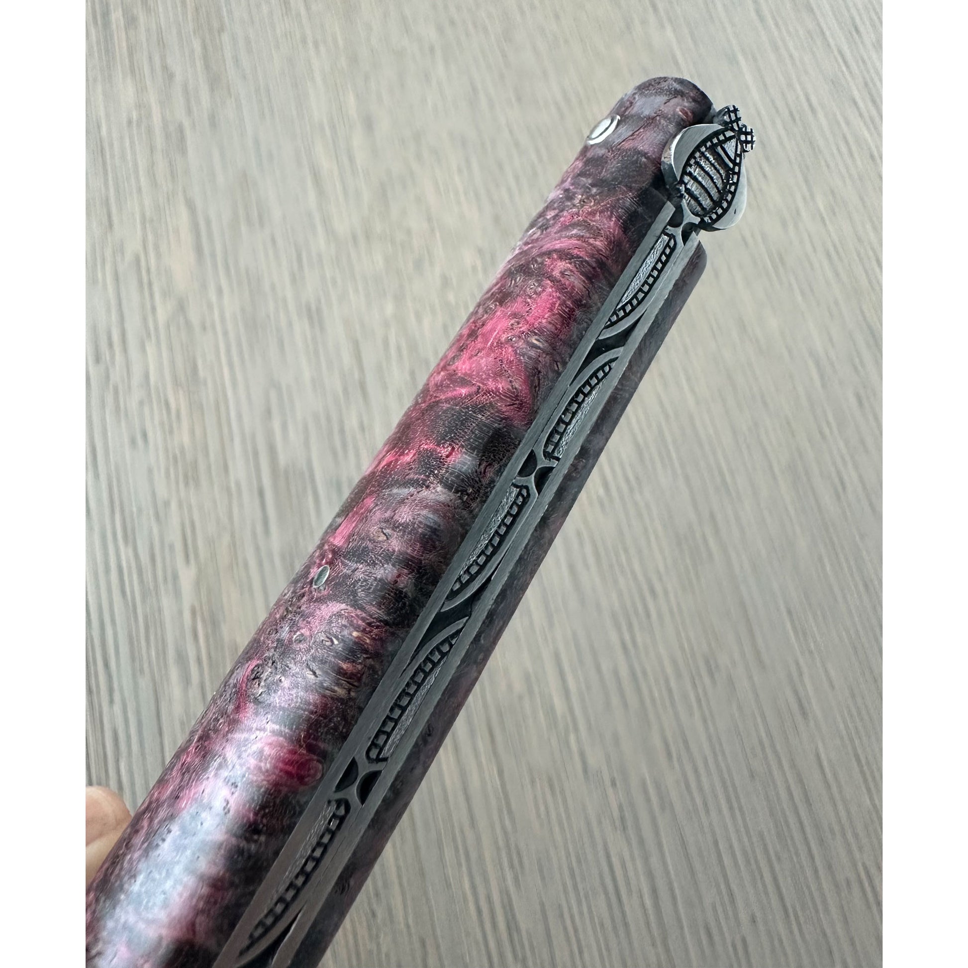 A close-up of a folding pocket knife with a marbled pink and black wood-grain handle, shown partially open against a gray wood-textured background.