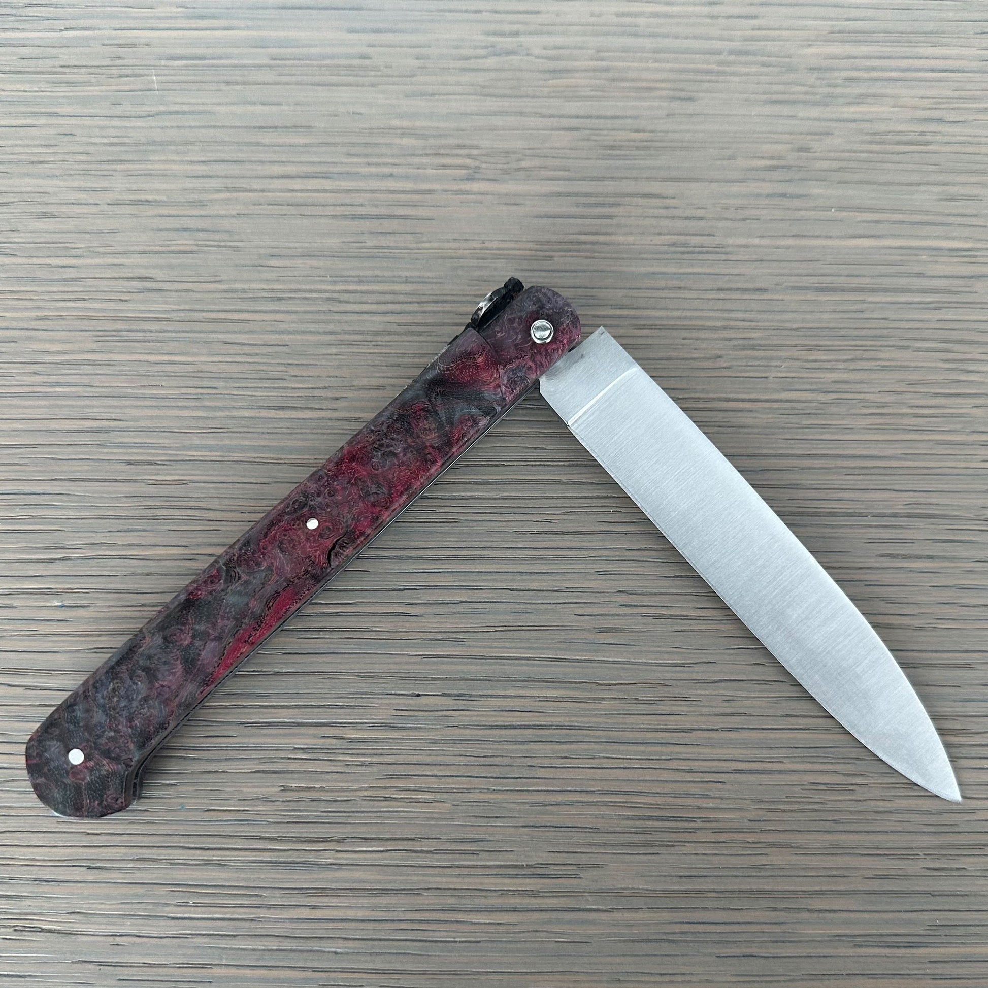 A folding pocket knife with a textured red and black handle and a satin-finished silver blade, shown open on a gray wood-grain surface.