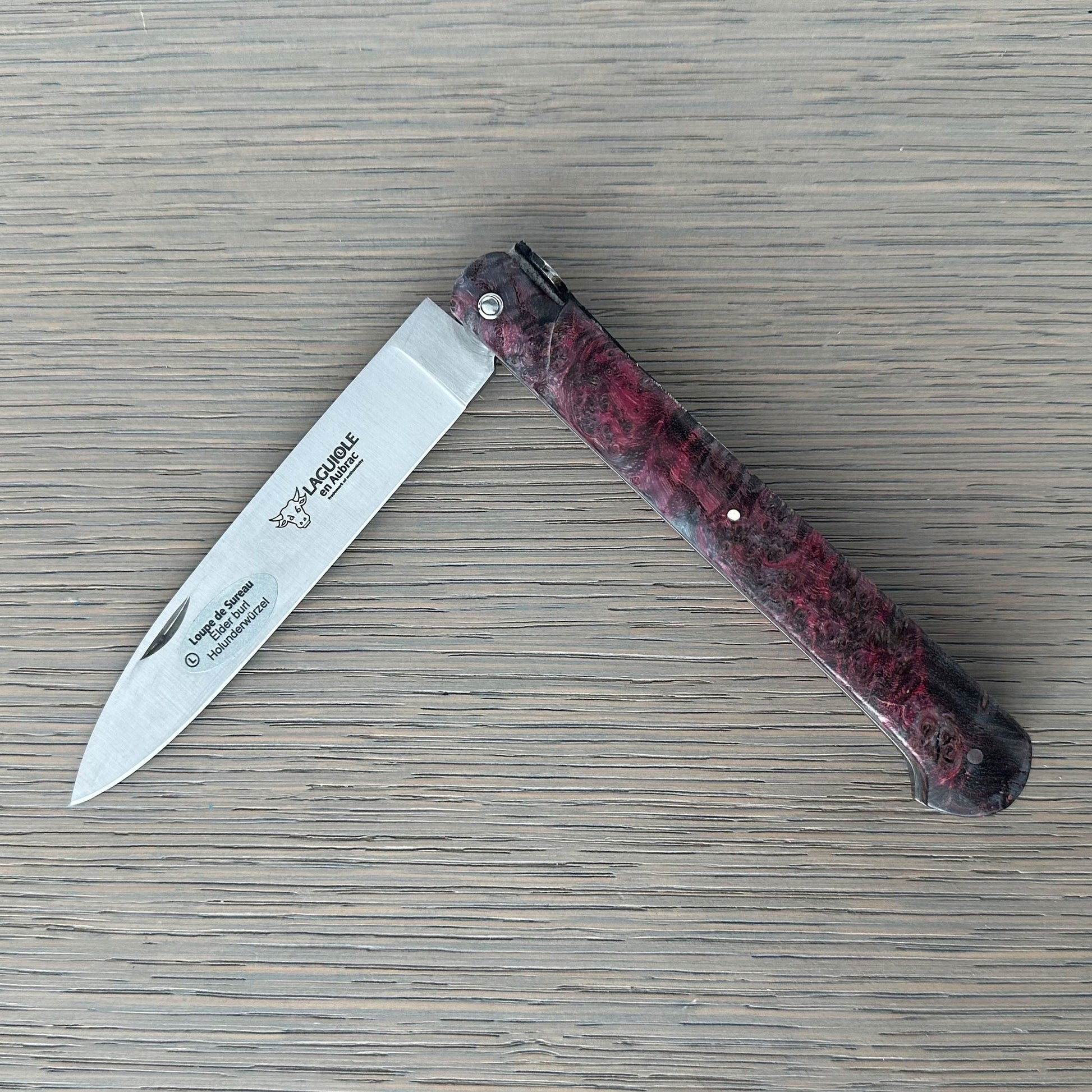 A folding pocket knife with a silver blade and a dark red and black marbled handle, shown open on a gray wood-grain surface. 