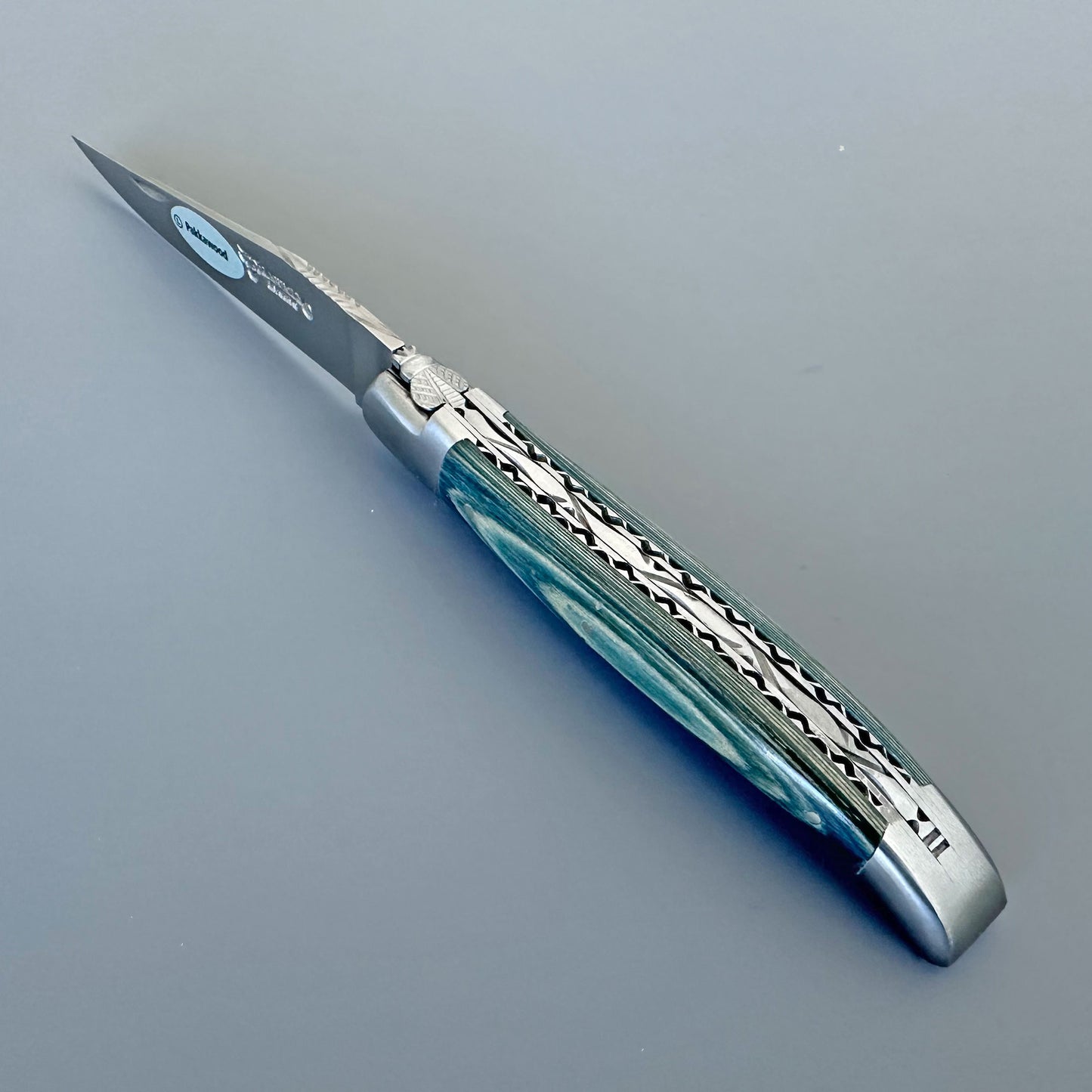 A folding pocket knife with a silver steel blade and a decorative handle featuring a blue tones marbled pattern with silver geometric accents, shown on a neutral gray background.