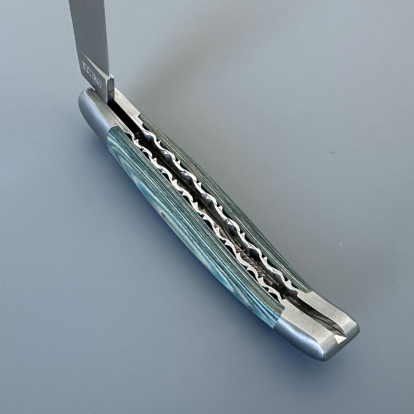 A close-up of a folding pocket knife with a silver metal blade and handle, featuring a wavy patterned inlay and light blue-green striped accents on a gray background.
