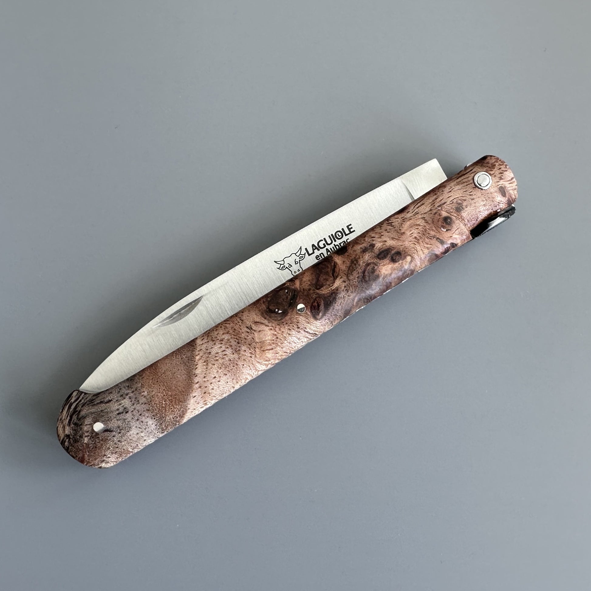 A folding pocket knife with a textured brown burl wood handle and a silver stainless steel blade, shown  closed. The brand name 'Laguiole en Aubrac is printed on the blade.