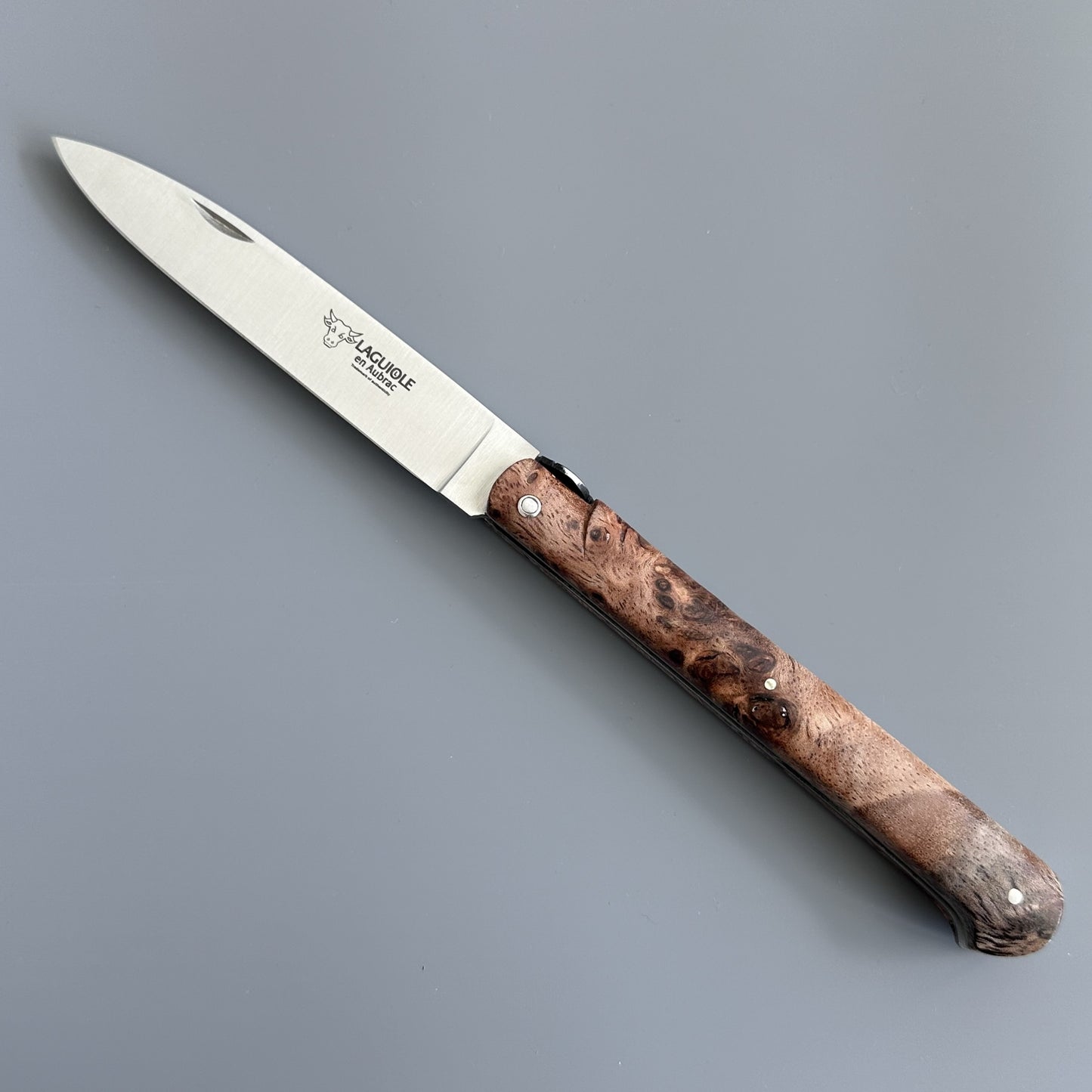 A folding-blade knife with a silver steel blade and and Albizia richly textured brown burl wood handle, shown diagonally on a neutral gray background. The blade features the Laguiole en Aubrac logo