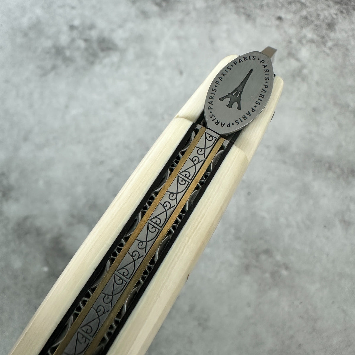 Laguiole en Aubrac close-up of a folding knife partially extended, showing a light natural wood handle with a black and gold patterned center stripe and a silver-toned oval metal end engraved with the Eiffel Tower and the words 'PARIS'.