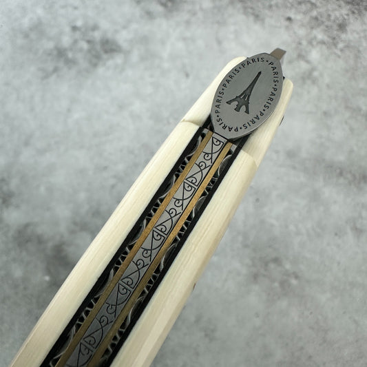 Laguiole en Aubrac close-up of a folding knife partially extended, showing a light natural wood handle with a black and gold patterned center stripe and a silver-toned oval metal end engraved with the Eiffel Tower and the words 'PARIS'.