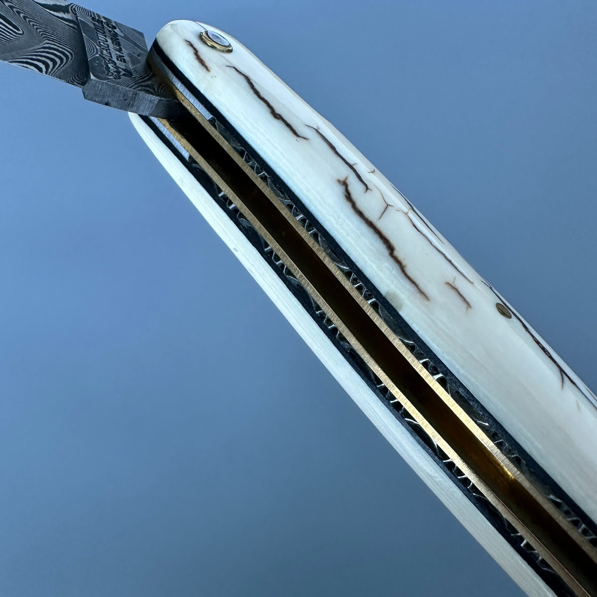 Laguiole en Aubrac close-up of a folding pocket knife with a Damascus steel blade partially extended, set against a solid blue background. 