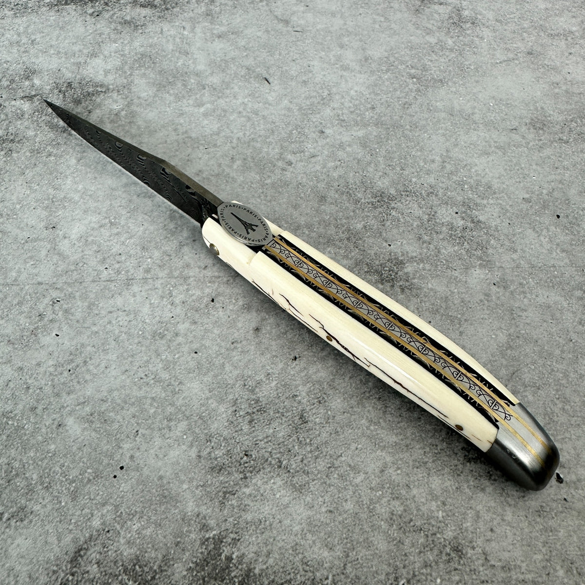 Laguiole en Aubrac folding pocket knife with a damascus blade and a mammoth ivory crust featuring black and gold inlay patterns, resting diagonally on a textured gray concrete surface.