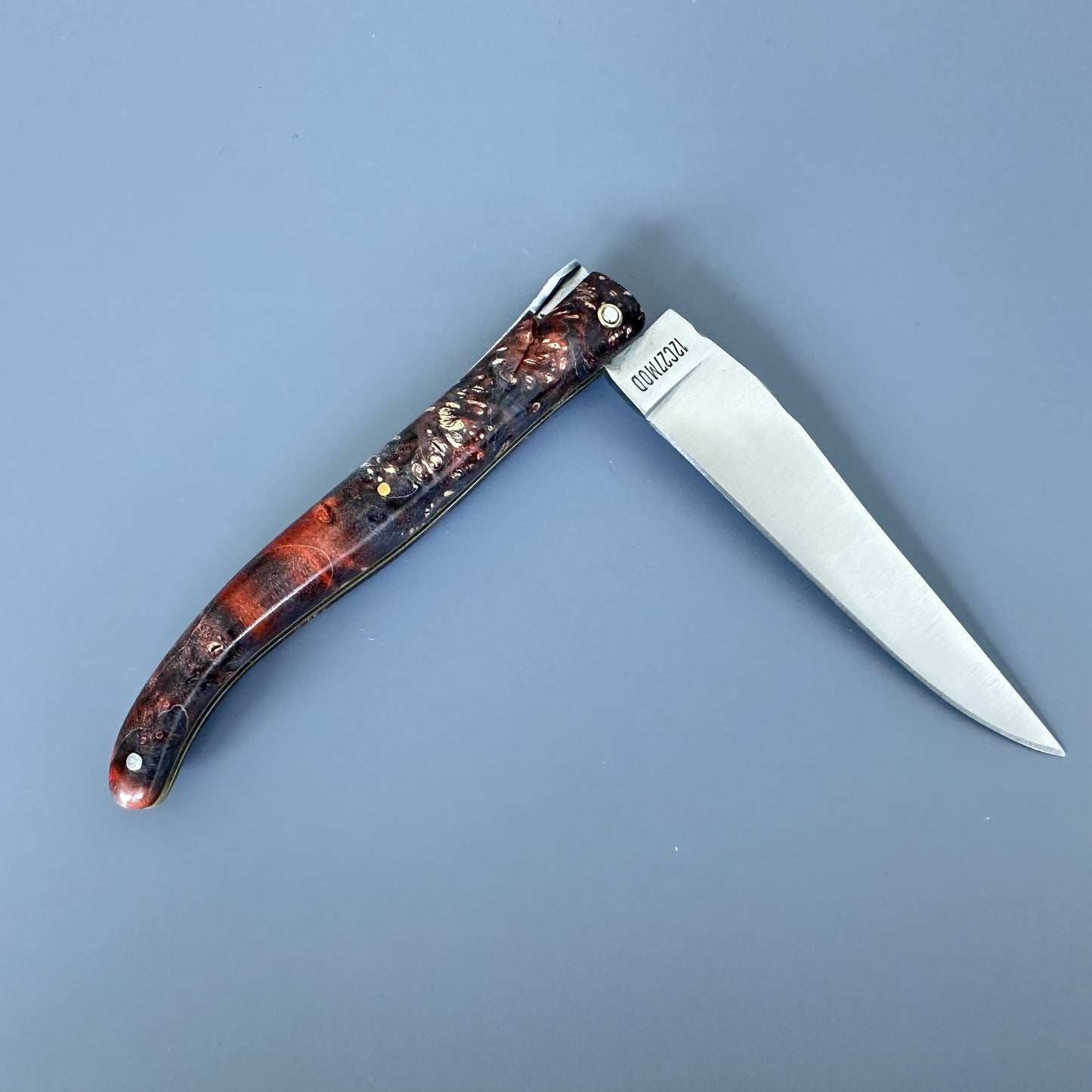 A folding pocket knife with a satin silver blade and a marbled reddish-brown and black handle, shown open on a solid slate-blue background. 