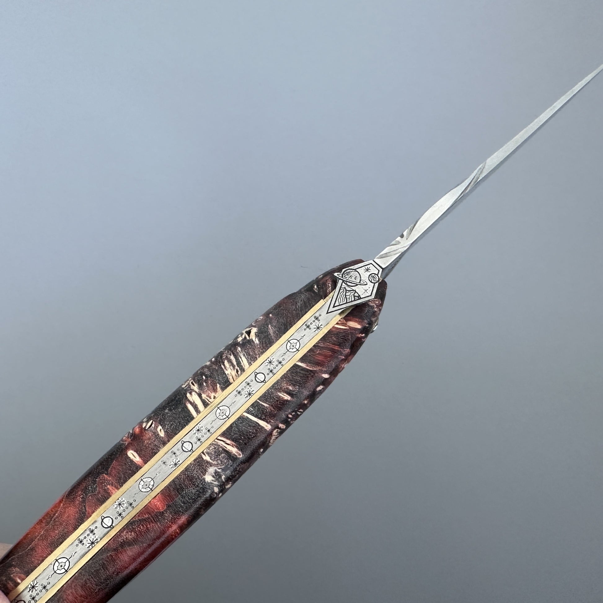 A close-up of a decorative folding pocket knife with a long, slender silver blade and an intricately carved handle featuring a reddish-brown wood grain, gold-toned metal inlay, and silver geometric accents.