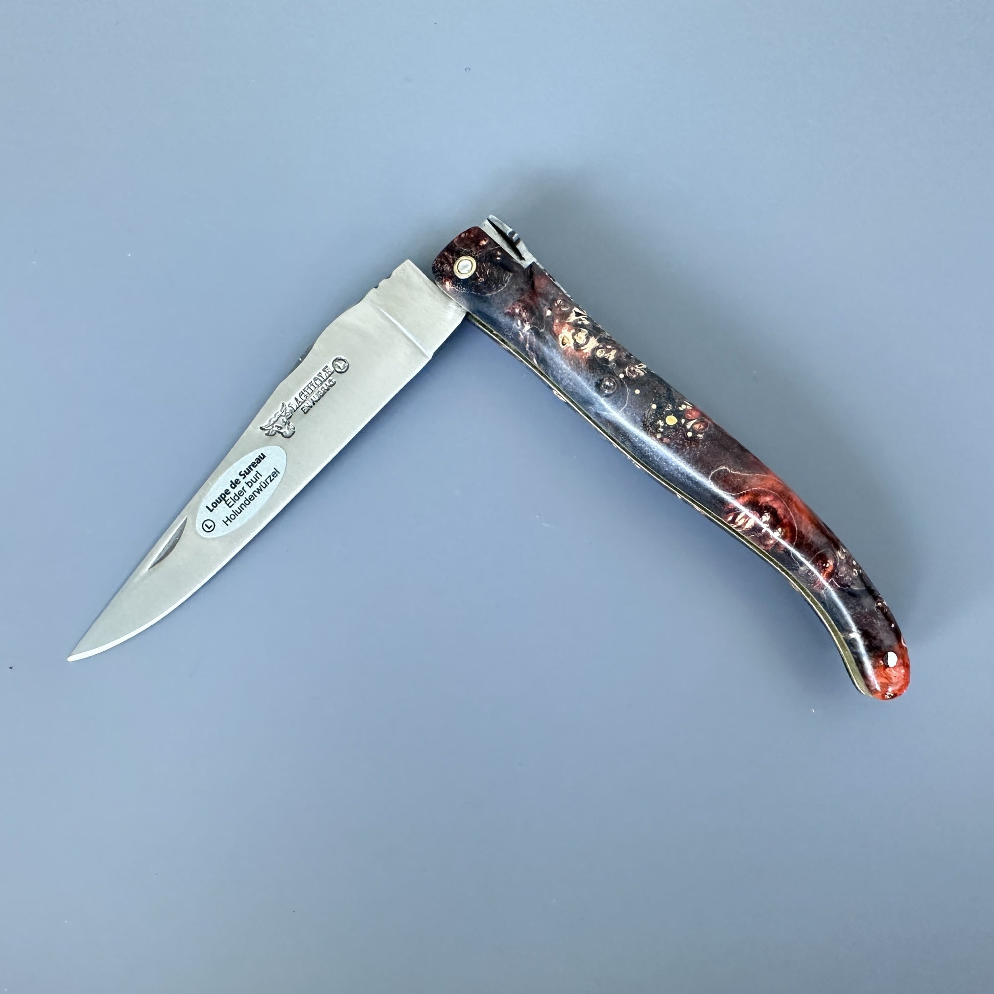 A folding pocket knife with a satin-finished silver blade and a marbled handle featuring black, red, and brown tones with subtle gold flecks, displayed on a solid blue-gray background.